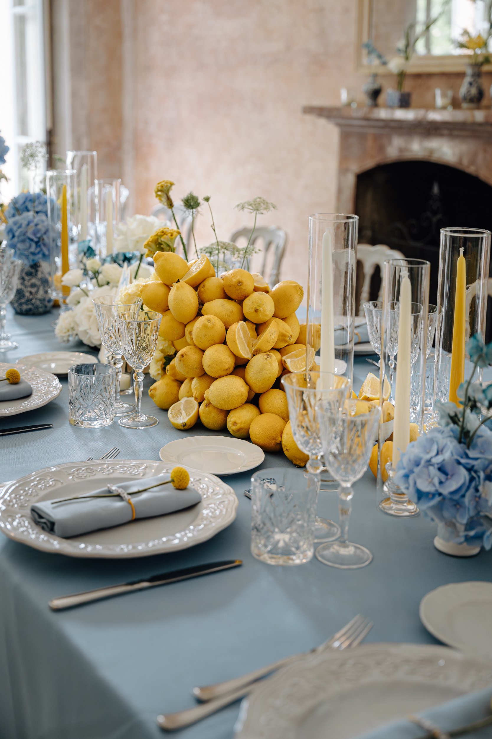 A table set for a celebration with a centerpiece of lemons and flowers, surrounded by glassware, plates, and cutlery, in a room with a fireplace and large windows.