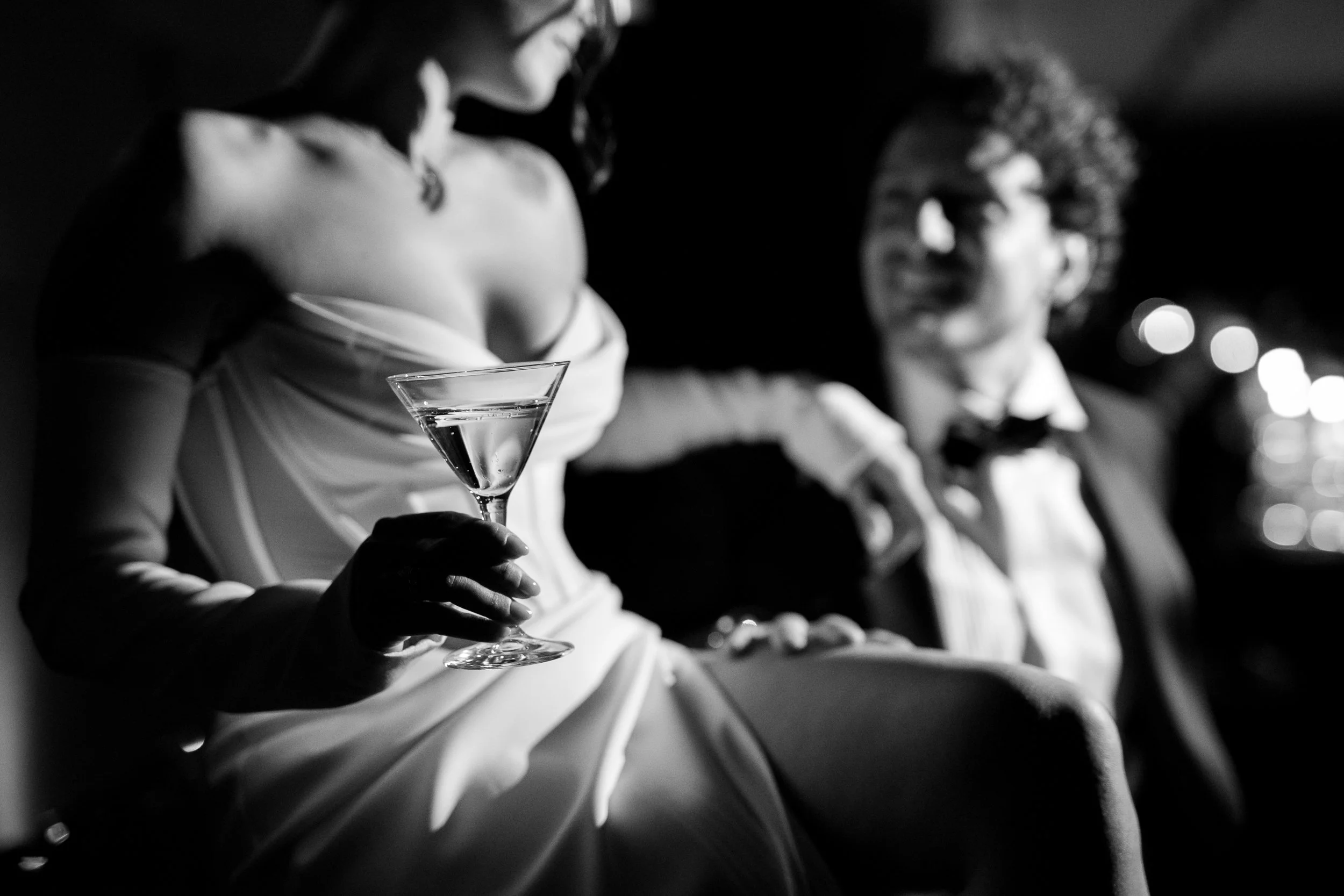 A woman in a white dress holding a cocktail glass, sitting with a man dressed as a tuxedo in a dimly lit setting.