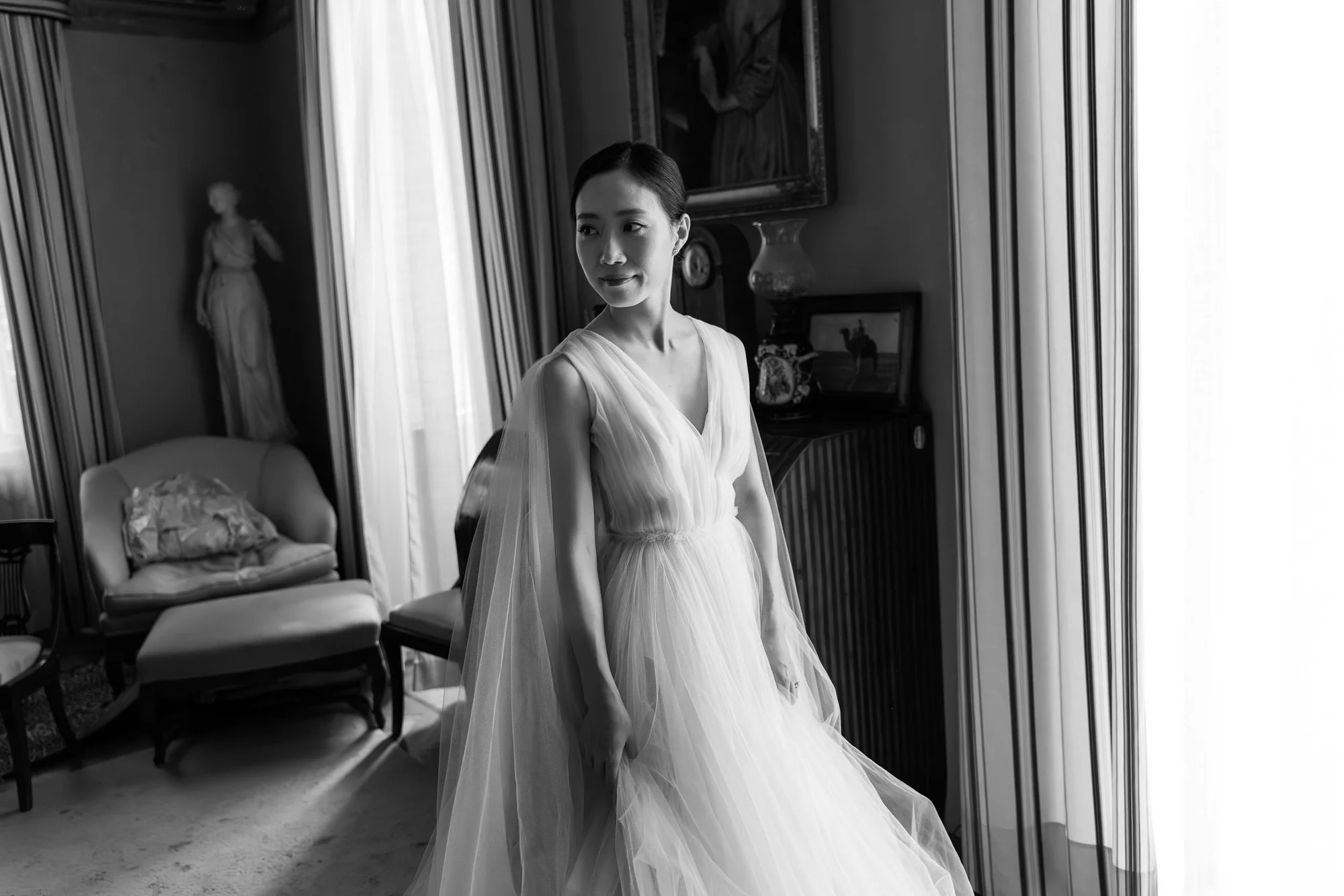 Black and white photo of a woman in a wedding dress standing in a room with classic decor, next to a large mirror and a chair.