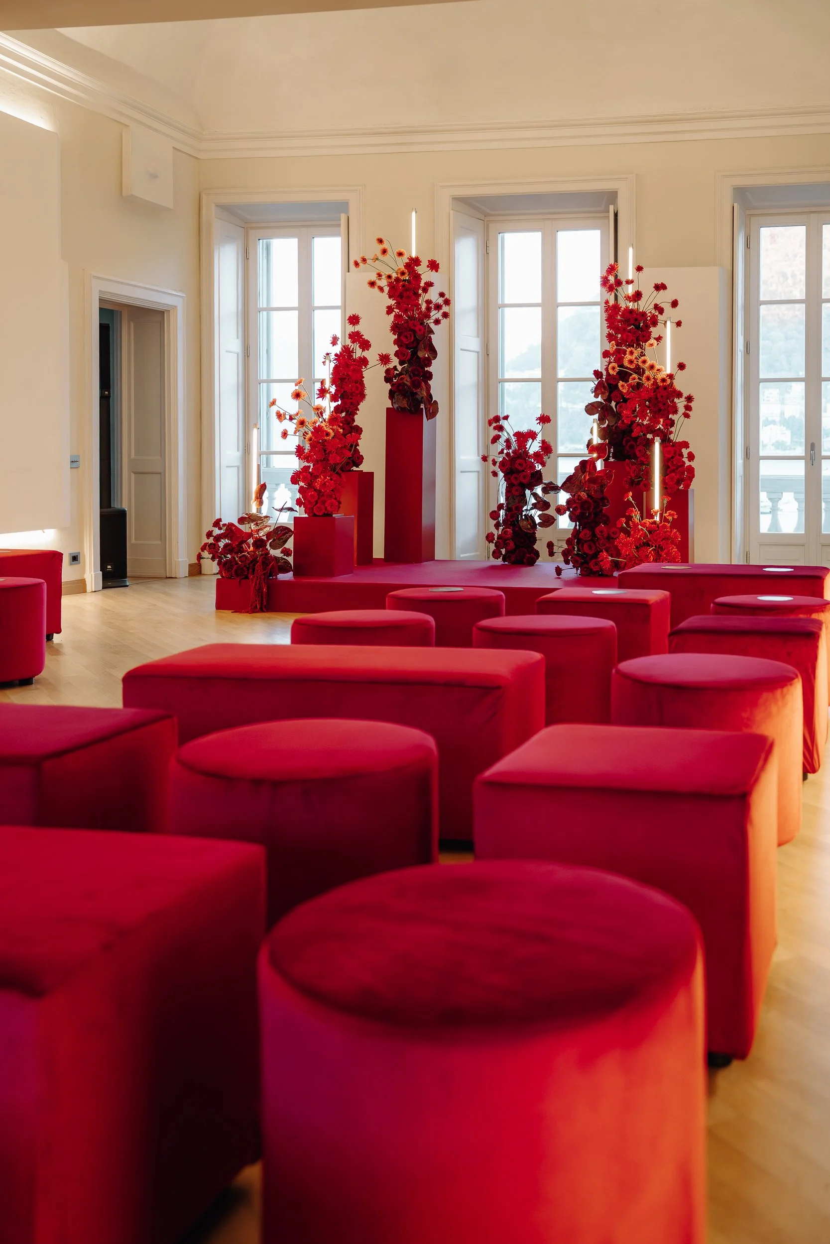 A room with red circular ottomans arranged in a circle or semicircle, and decorated with tall red floral arrangements on red pedestals, placed in front of large windows with white frames, letting in natural light.