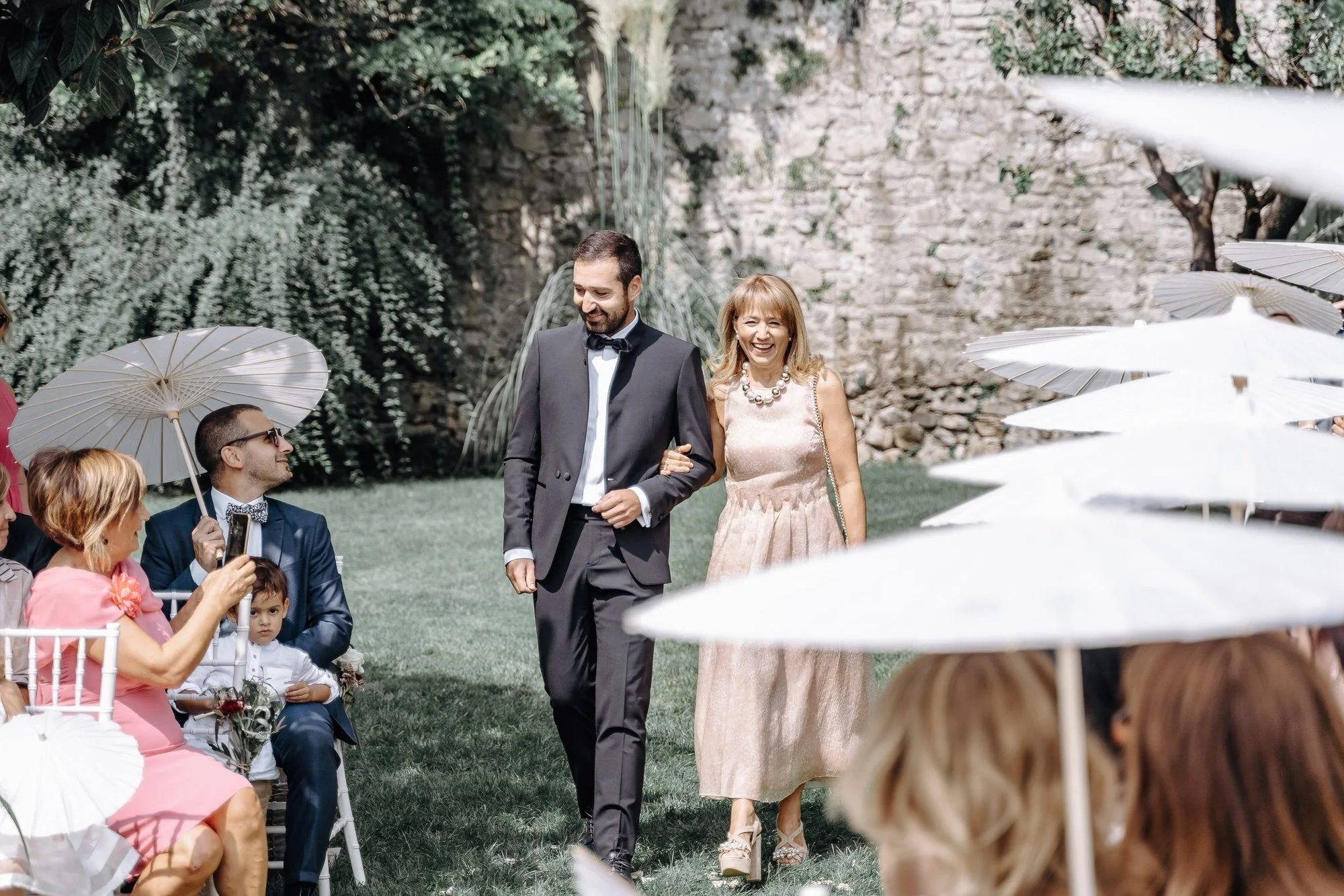 A wedding outdoor ceremony with a groom in a tuxedo walking with a woman in a light-colored dress, surrounded by seated guests holding parasols, on a grassy area with stone walls and green foliage in the background.