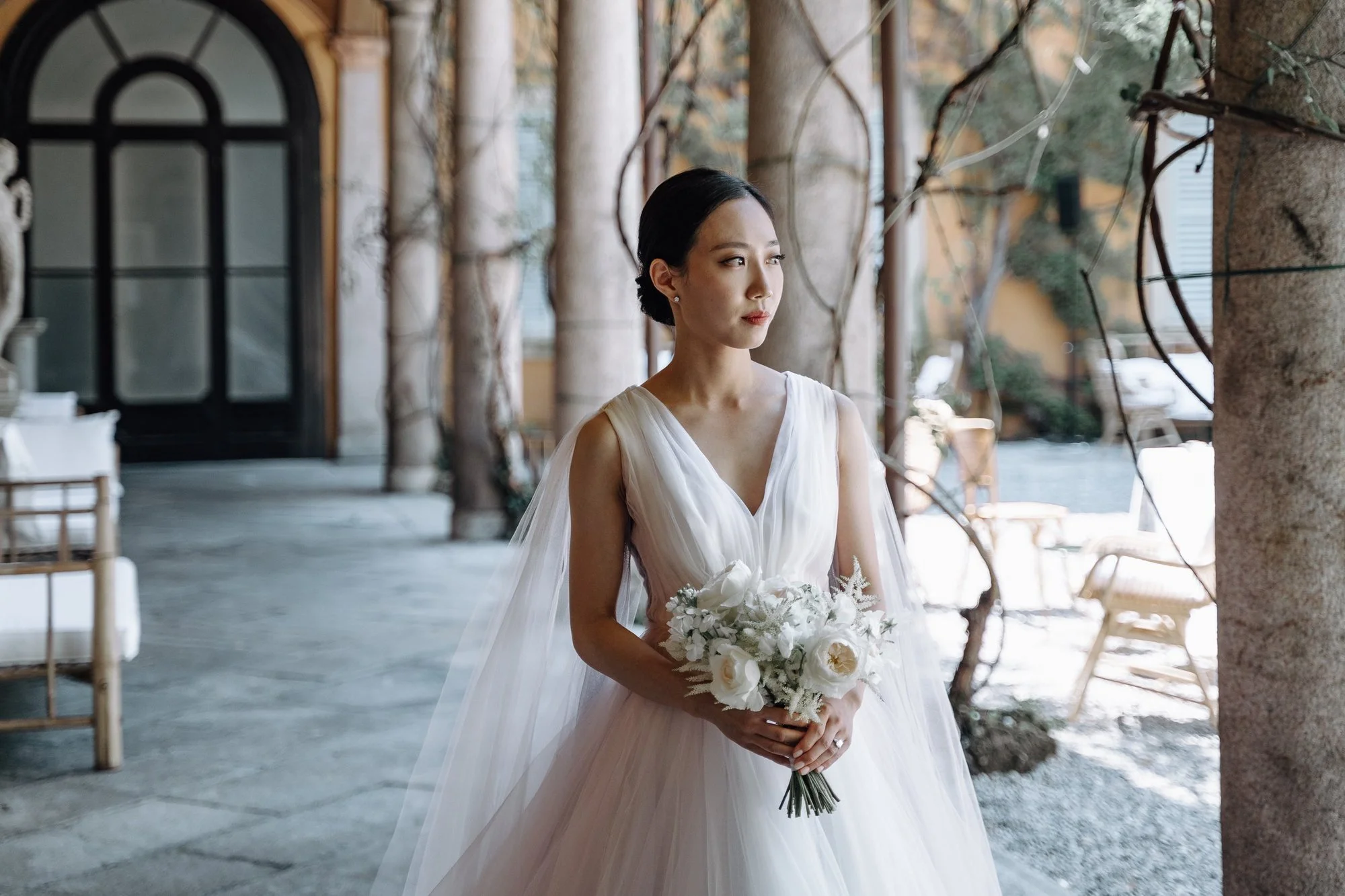 A woman in a white wedding dress holding a bouquet of white flowers, standing outdoors near stone columns and trees.