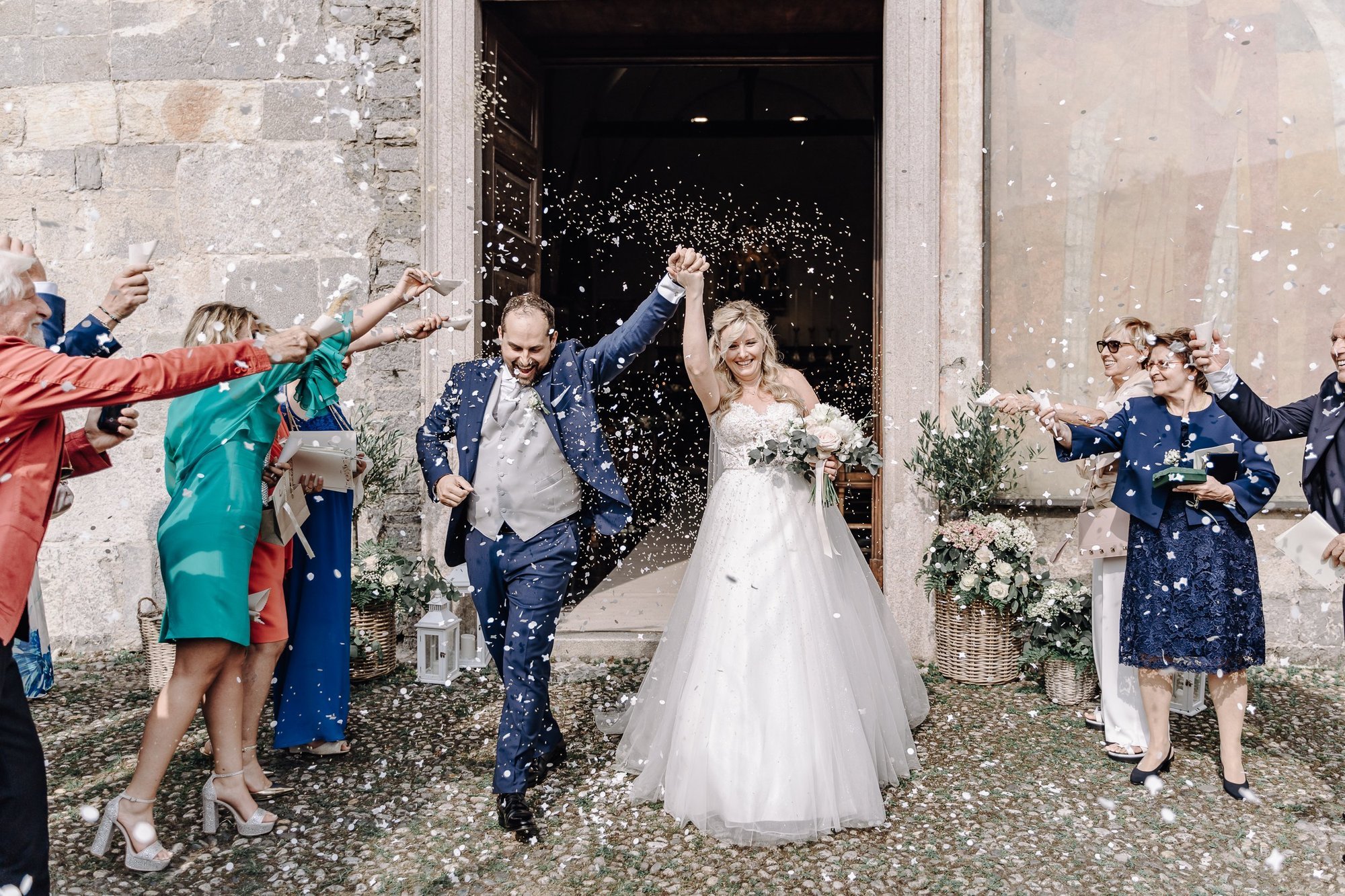 A bride and groom exit a wedding ceremony as guests throw confetti in the air. The bride is wearing a white wedding dress and holding a bouquet, and the groom is in a blue suit. Everyone is smiling and celebrating in front of a stone building.