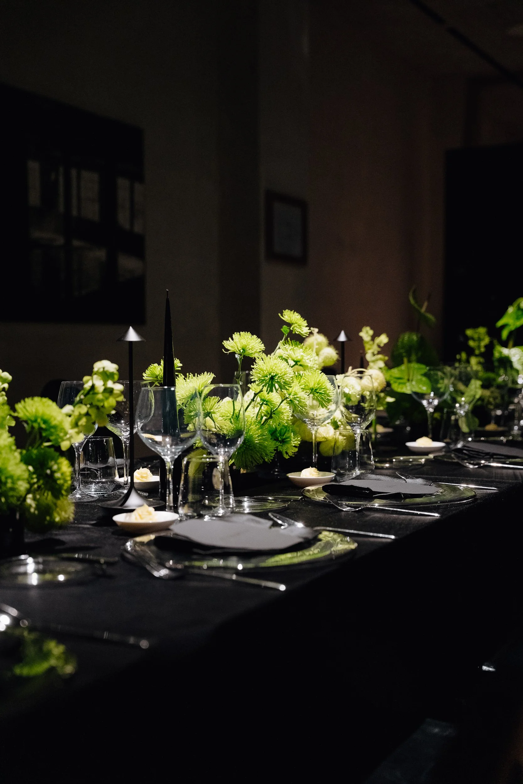 A darkly lit dining table decorated with green flowers, wine glasses, and small bowls with butter. The background shows blurred framed pictures on the wall.