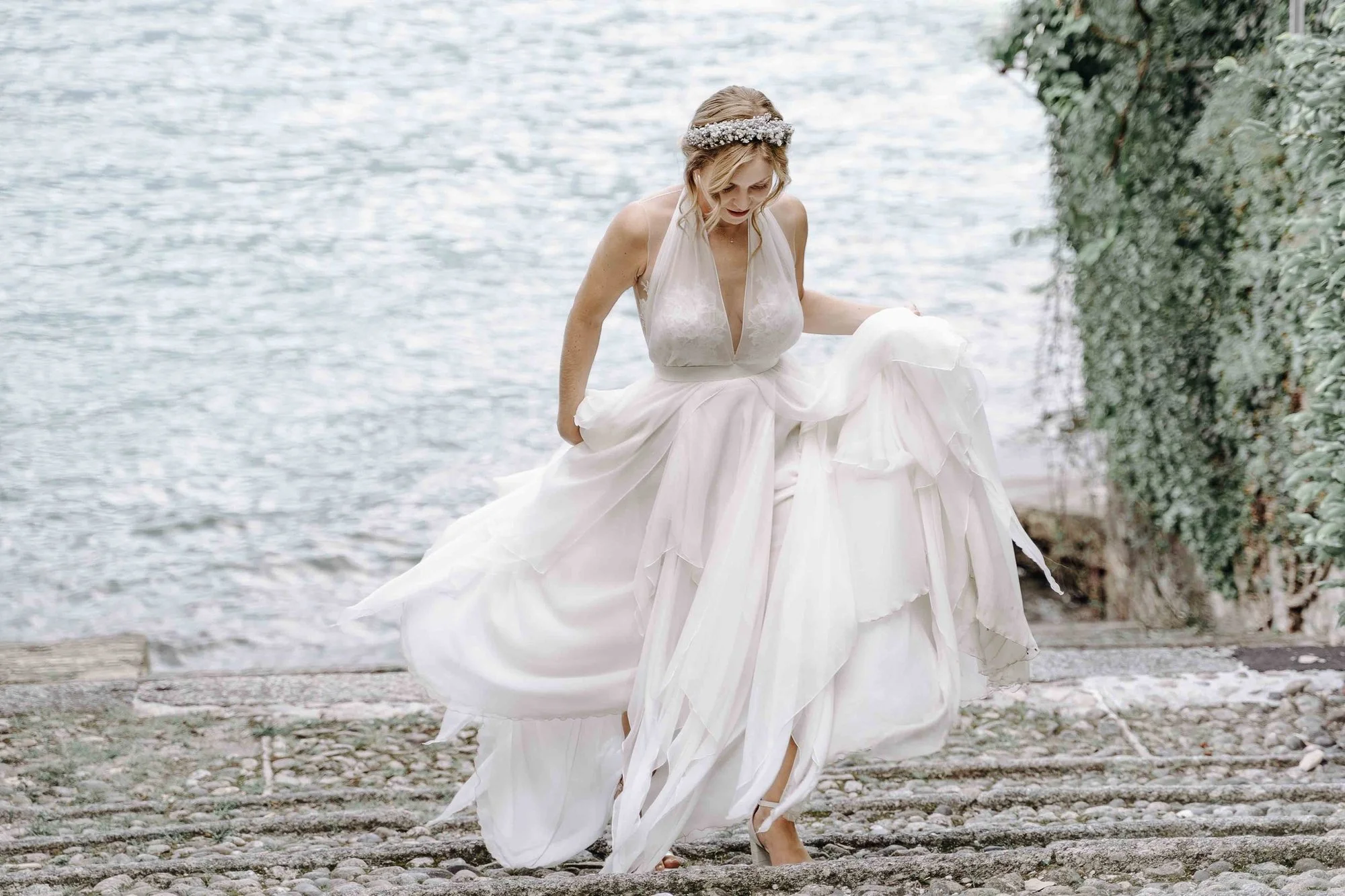 A woman in a white wedding dress walking along a pebbled shoreline by a body of water, with lips slightly parted and looking down, with greenery on the right side.