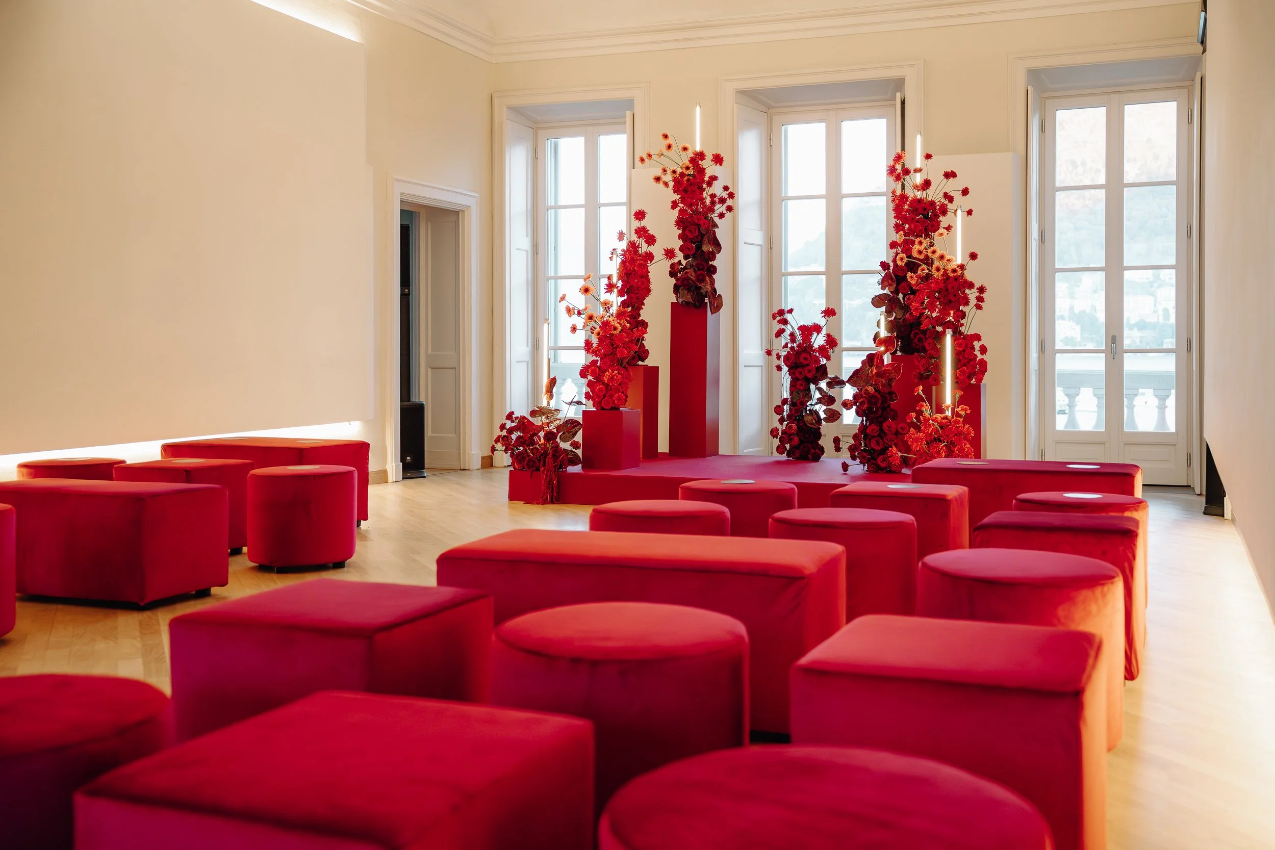 A spacious room with large windows, decorated with tall red floral arrangements and red cushioned seating.
