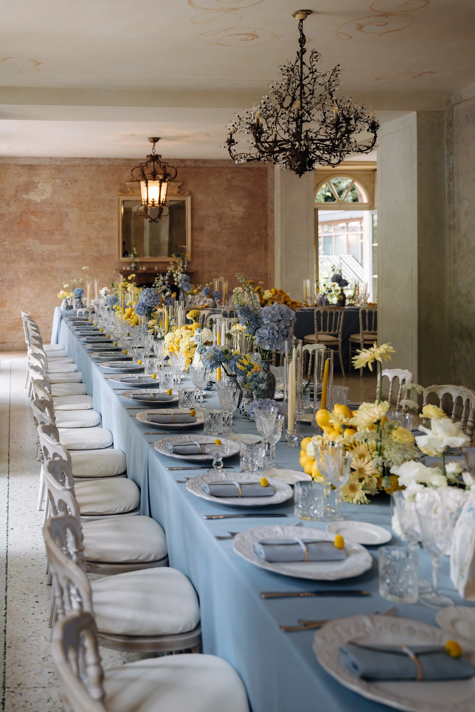 Long dining table covered with a light blue tablecloth, decorated with floral arrangements, candles, and table settings, in a spacious indoor banquet hall with vintage chandeliers and large window letting in natural light.