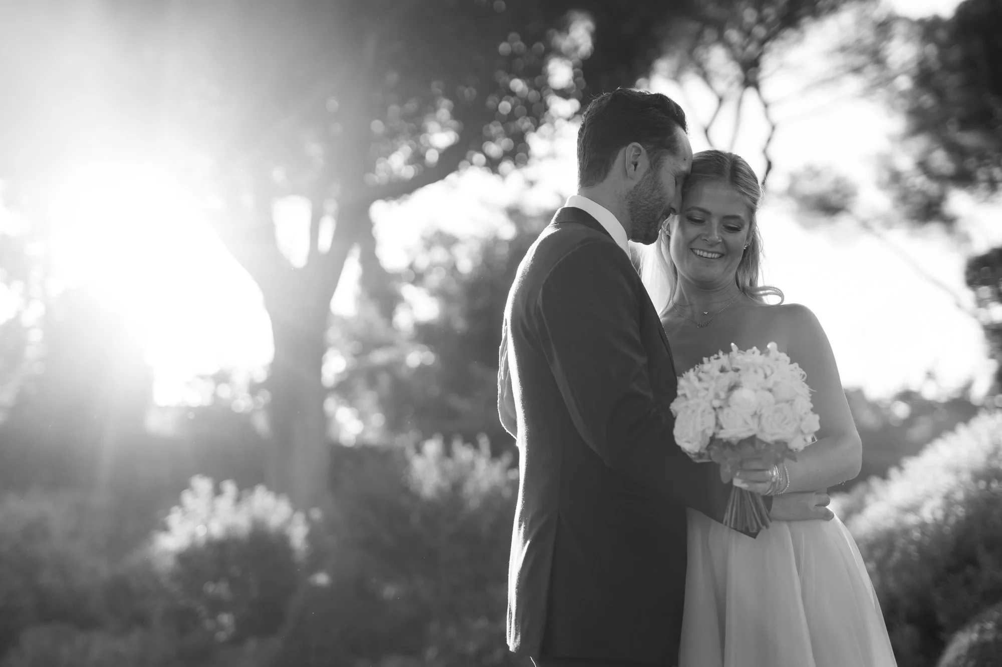 A black and white photo of a bride and groom embracing outdoors during a wedding, with the groom kissing the bride's forehead and the bride smiling, holding a bouquet of flowers, with trees and sunlight in the background.