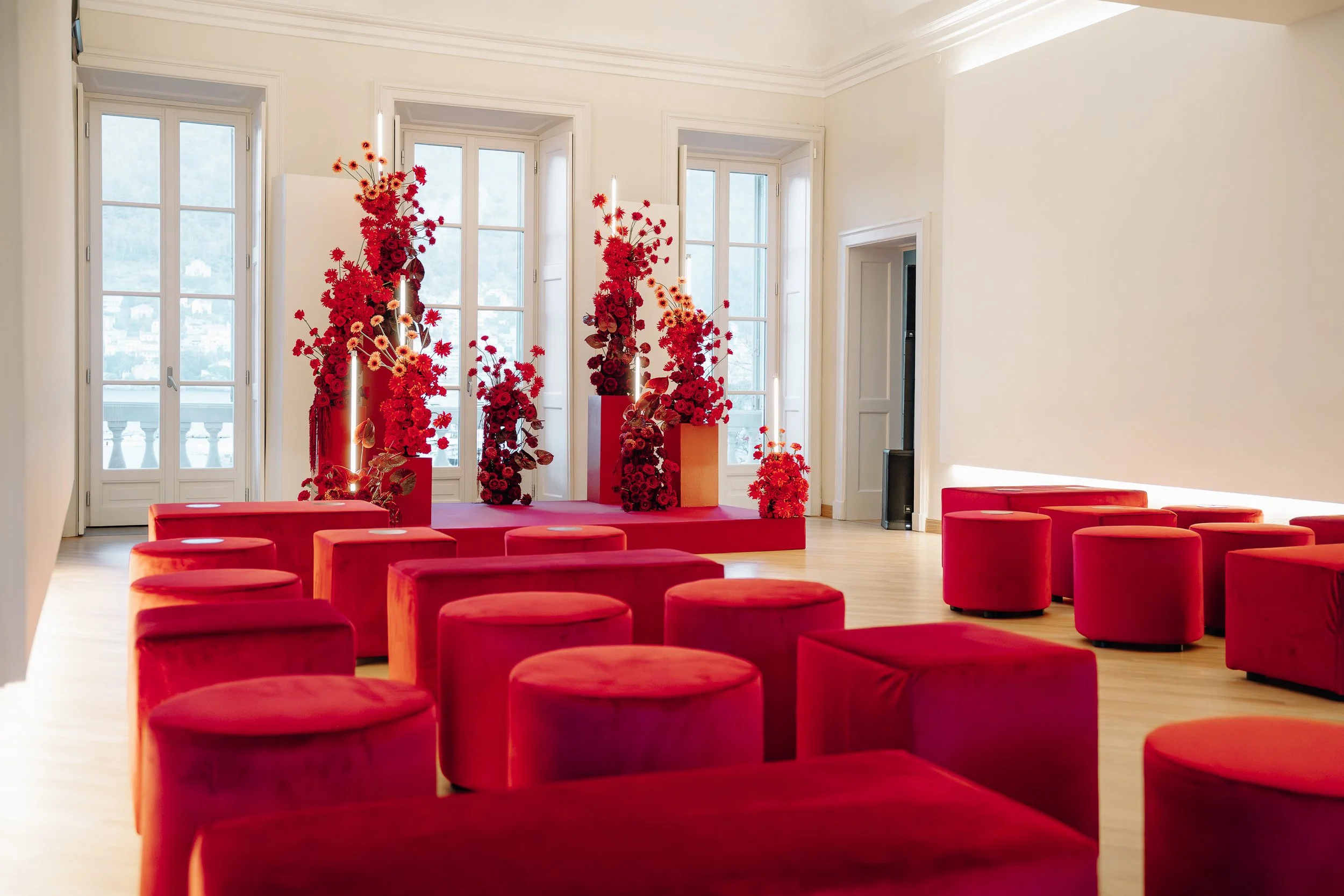 An event space with multiple red velvet upholstered stools arranged in rows, a stage decorated with large red floral arrangements, and tall white windows letting in natural light.
