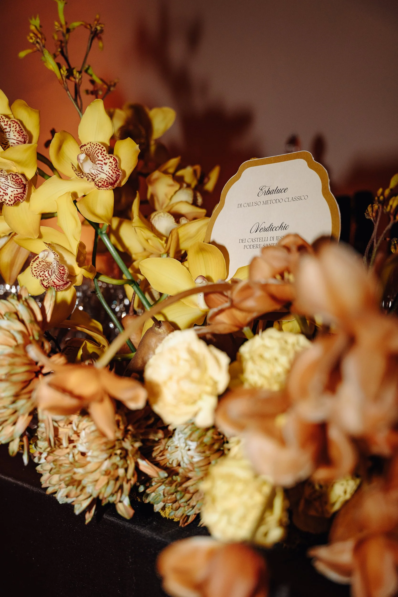 Close-up of yellow orchids with red and white patterned centers surrounded by orange and brown flowers, with a partially visible card in the background.