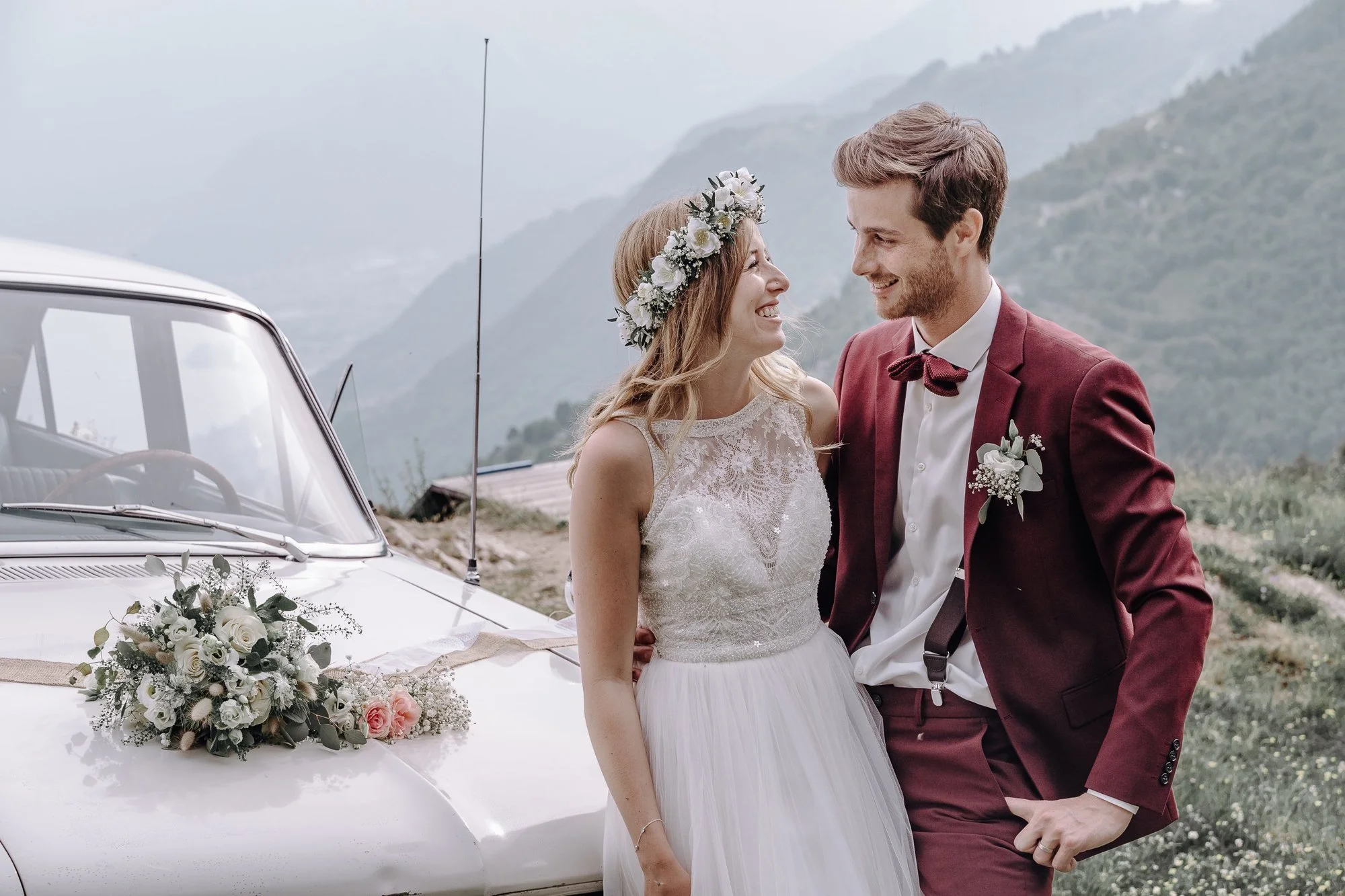 A bride and groom are smiling at each other outdoors, standing next to a vintage white car decorated with flowers, with a scenic mountainous background.
