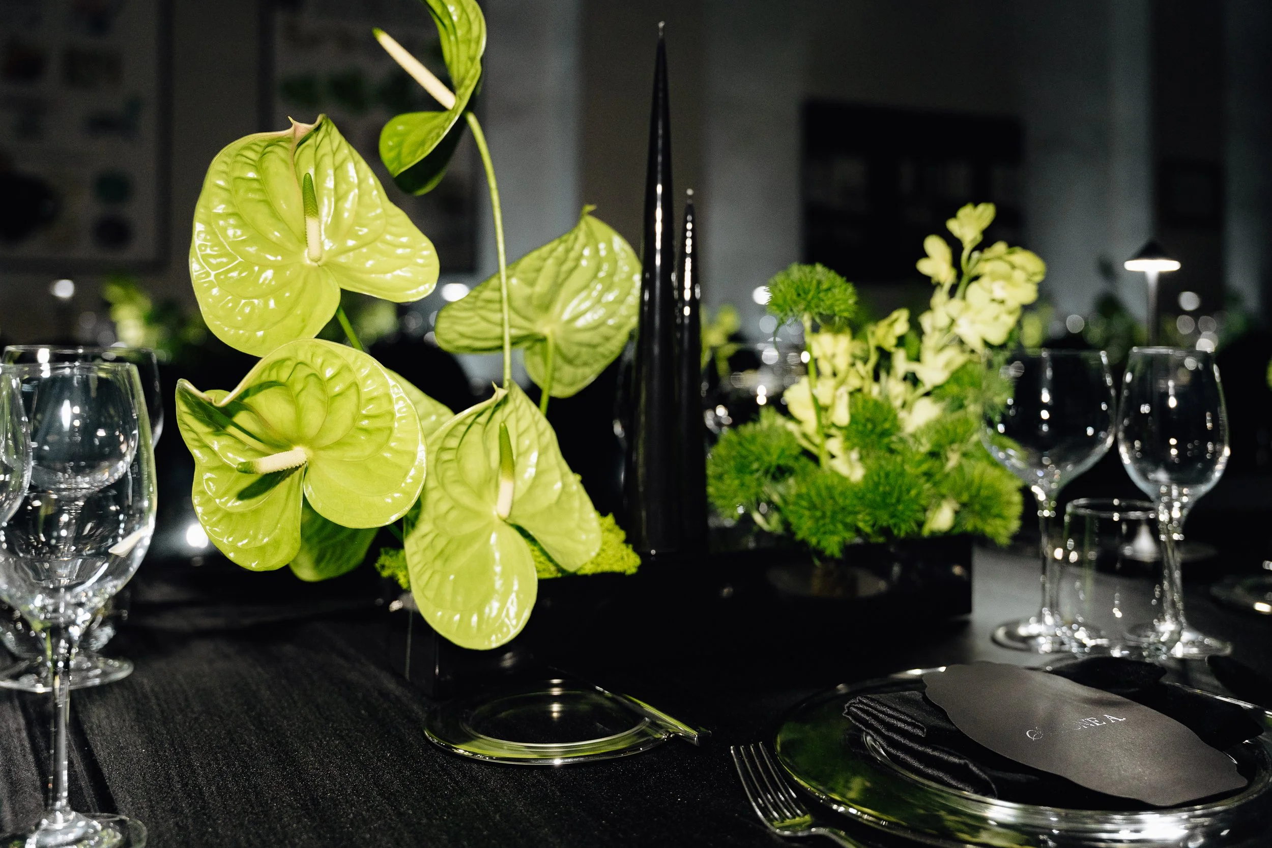 A close-up of a table setting with green floral arrangements including anthuriums, green leaves, and other greenery. Wine glasses, cutlery, and black placemats are also visible, suggesting an elegant dining setup.