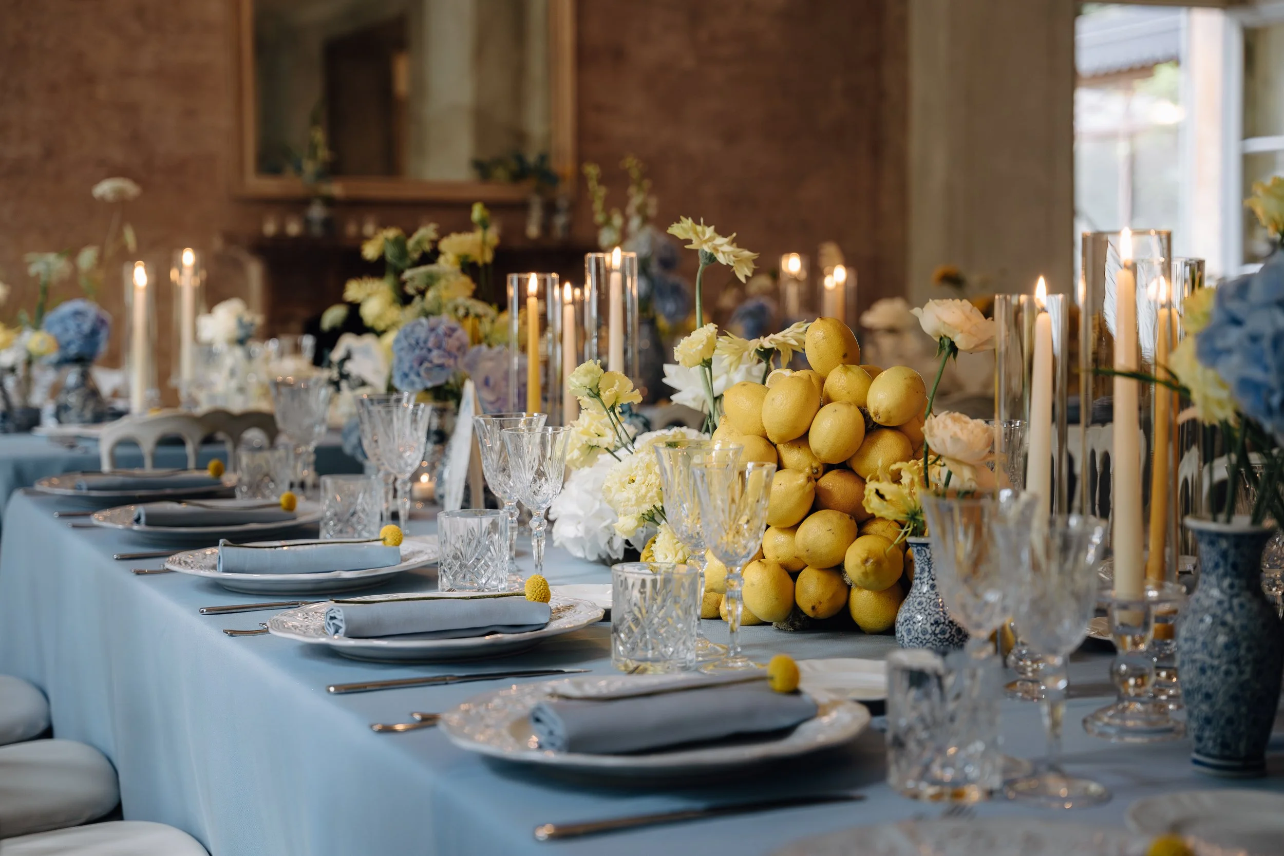 A decorated banquet table set for a special event, featuring glassware, plates with napkins, and a centerpiece of lemons, white and yellow flowers, with lit candles, in a rustic interior with large windows.