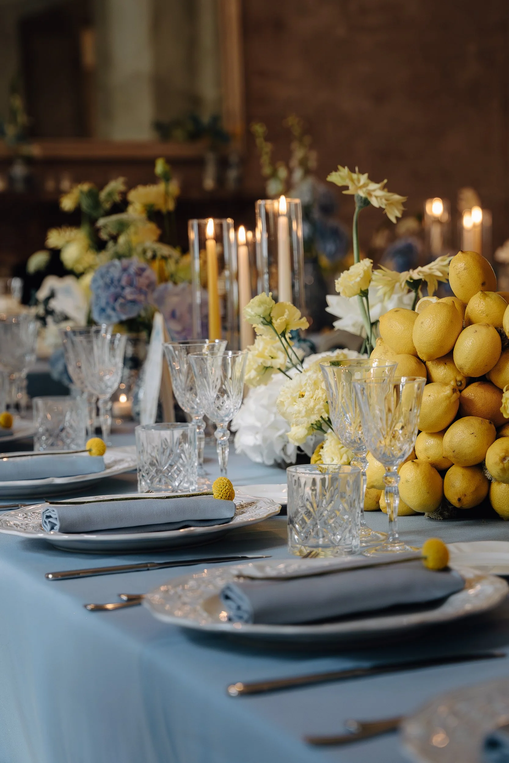 Elegant dining table set with crystal glassware, napkins, and cutlery, decorated with yellow lemons, white flowers, and candles in a warmly lit room.