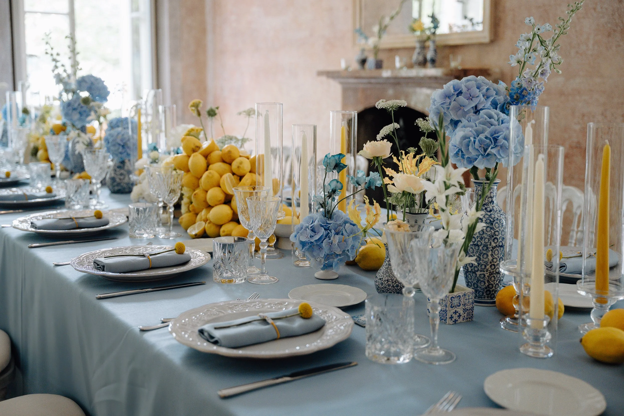 A beautifully decorated dining table with blue, yellow, and white floral arrangements, lemons, and elegant glassware for a celebration or special occasion in a bright room.