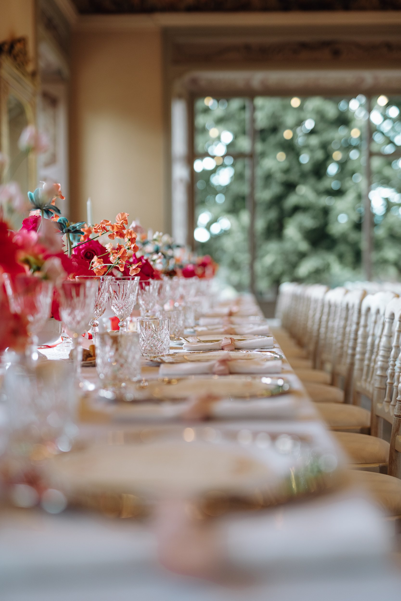 Elegant dining room table set with floral centerpieces, crystal glassware, and neatly folded napkins, with a large window showing green trees outside.