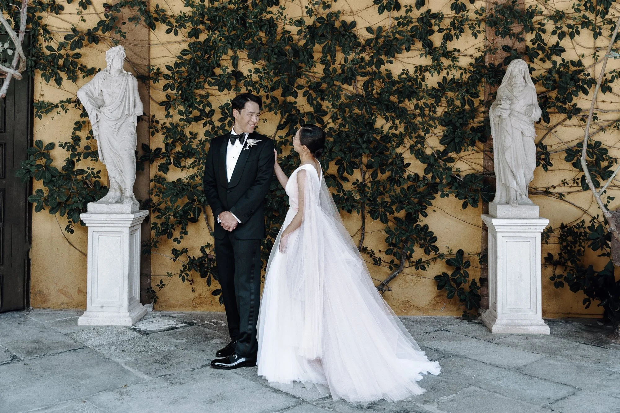A newlywed couple stands in front of a yellow wall covered in green ivy, flanked by two white statues on pedestals. The groom wears a black tuxedo with a bow tie, and the bride wears a white wedding gown with a long train. They smile at each other.