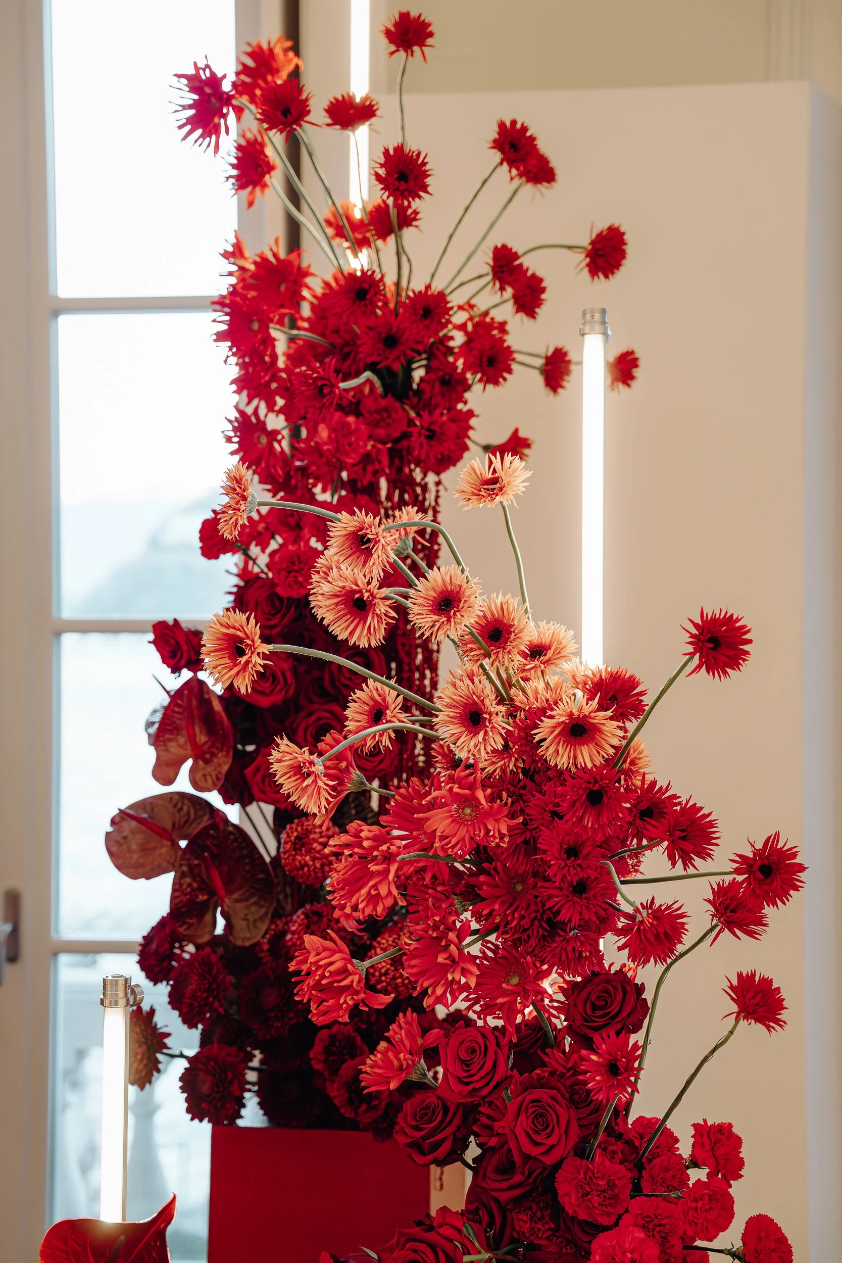 Tall flower arrangement with various red flowers, including roses and daisies, in a red container near a window with natural light
