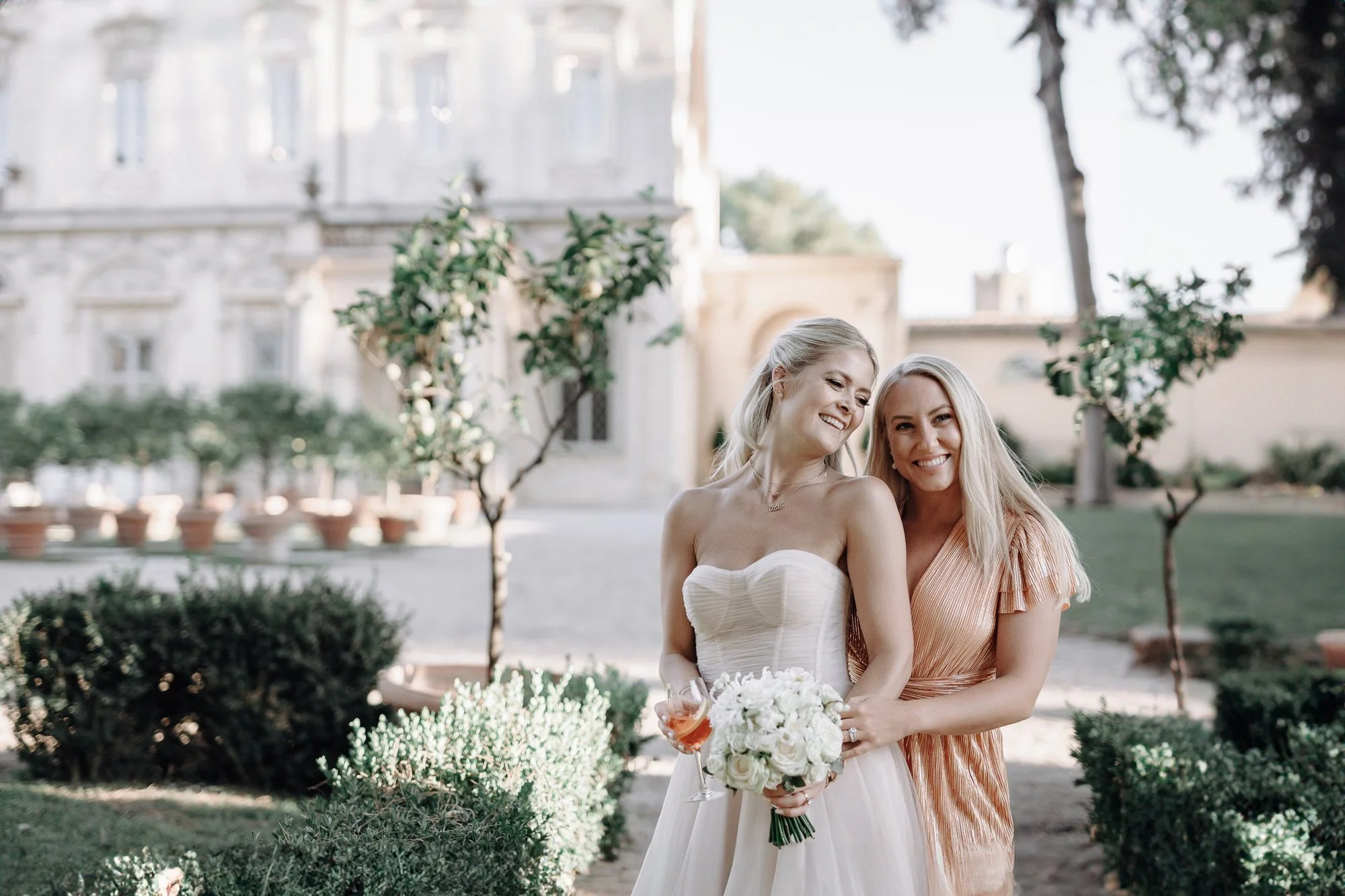 Two women, one in a white wedding dress holding a bouquet of white roses and a glass of wine, and the other in an orange dress, smiling and standing outdoors in a garden with trees and an elegant building in the background.