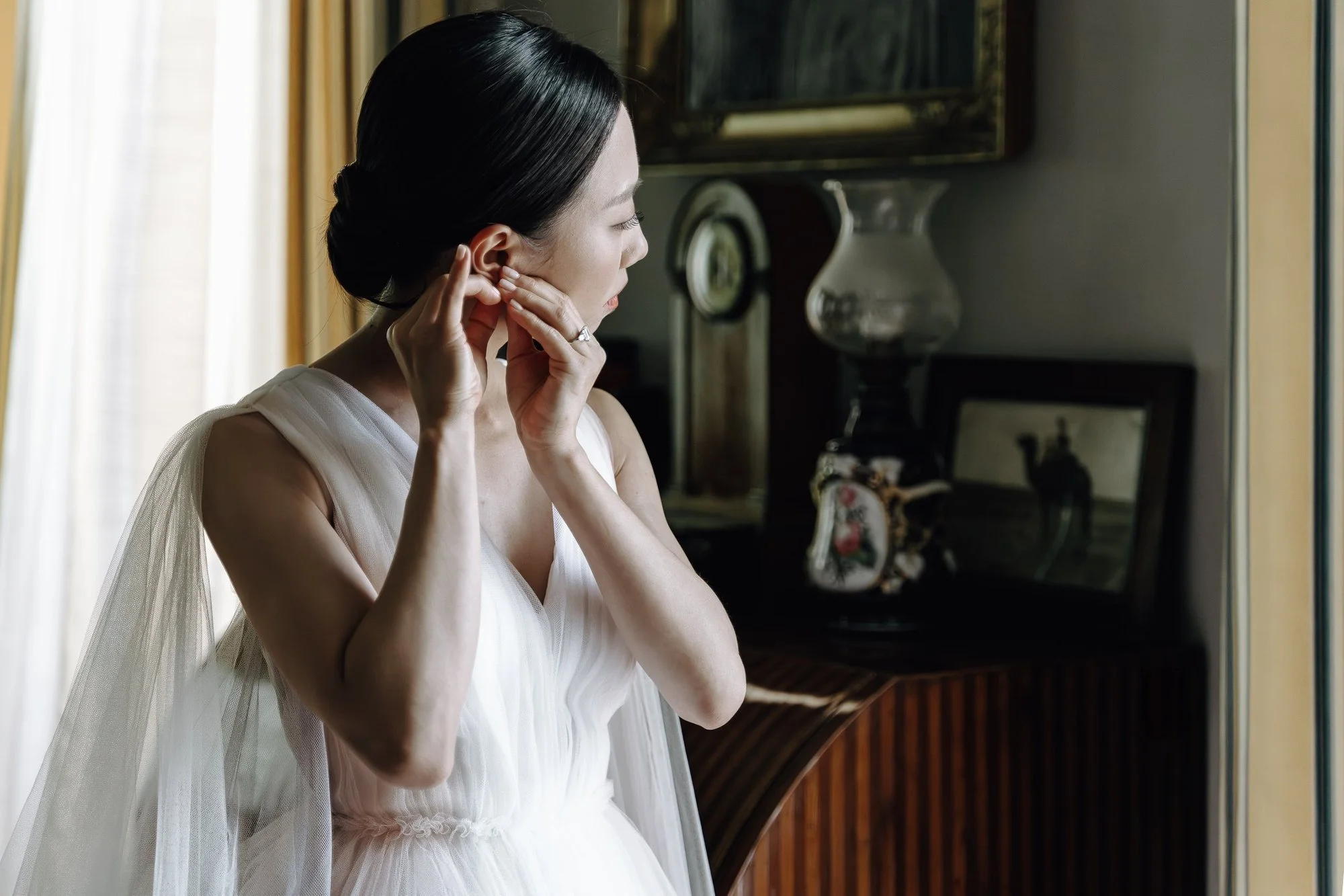 A woman in a white dress with dark hair styled in an updo is putting on an earring, standing indoors near a window with light-colored curtains. The background includes a vintage glass vase, a wall-mounted mirror, and other decorative items.