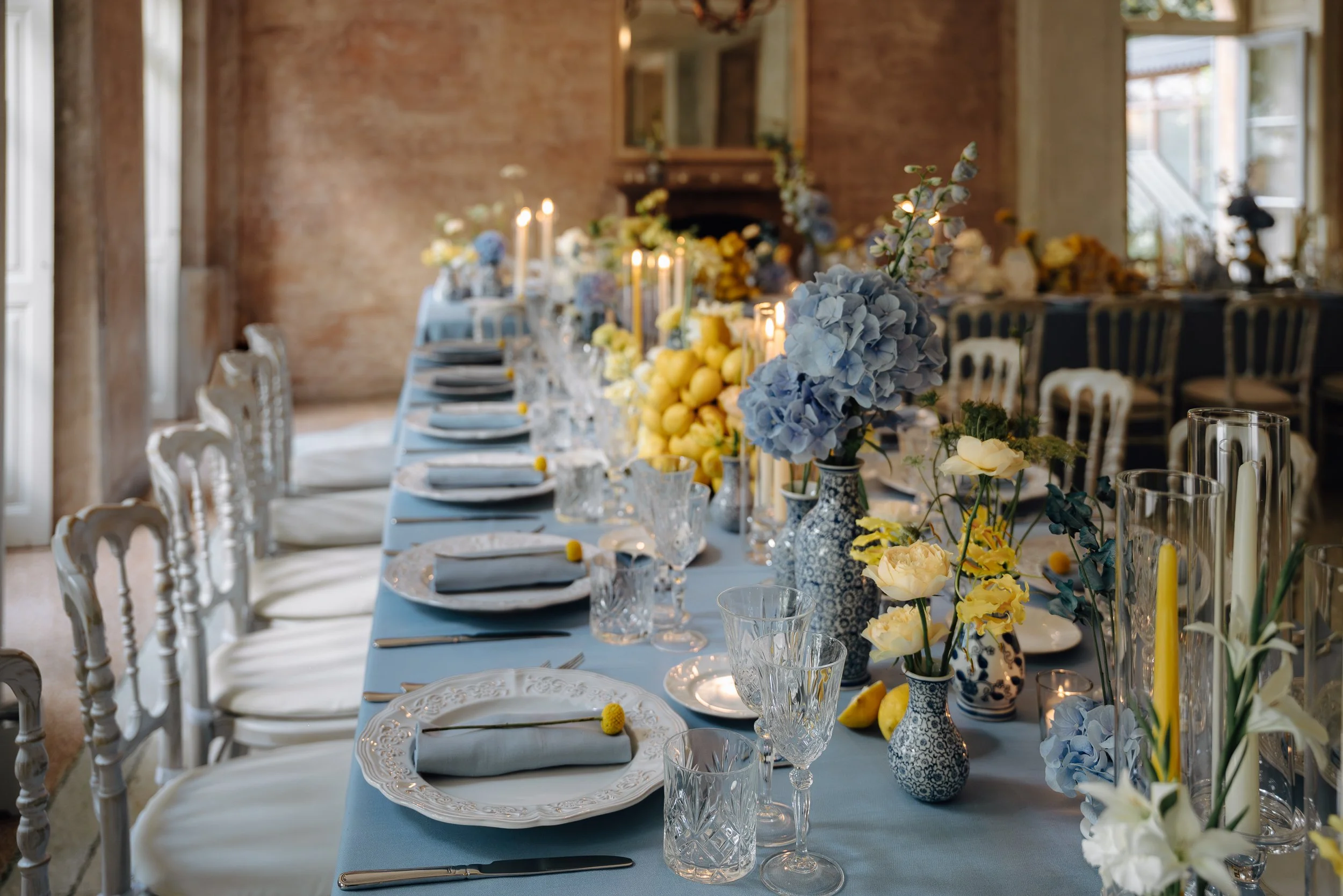 A long dining table set for a formal event with floral centerpieces, candles, glassware, and plates, in a room with brick walls and large windows.