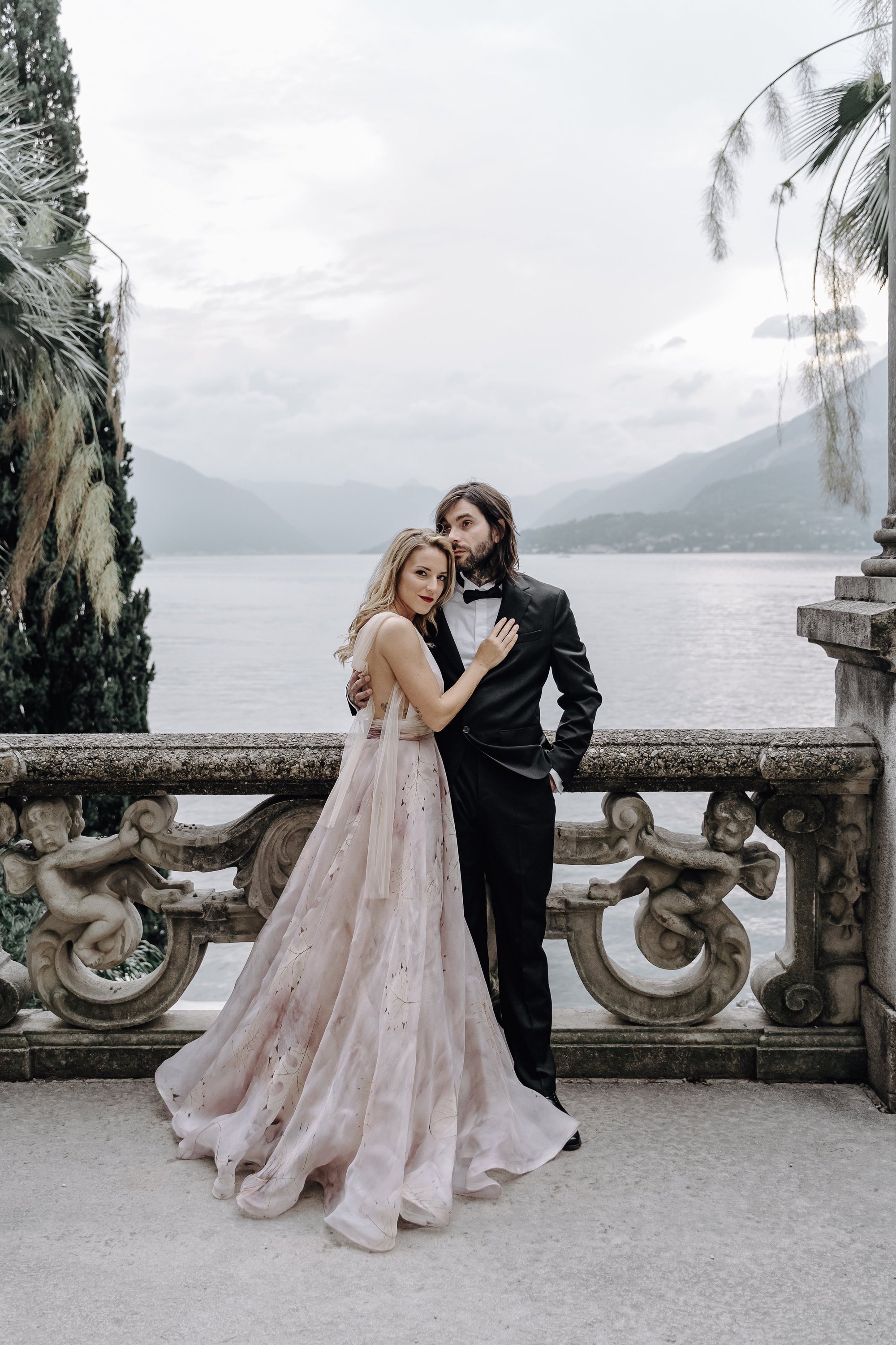 A couple dressed in formal attire, standing on a stone balcony with ornate cherub carvings, overlooking a lake with mountains in the background. The woman is in a flowing pink gown, and the man is in a black tuxedo.