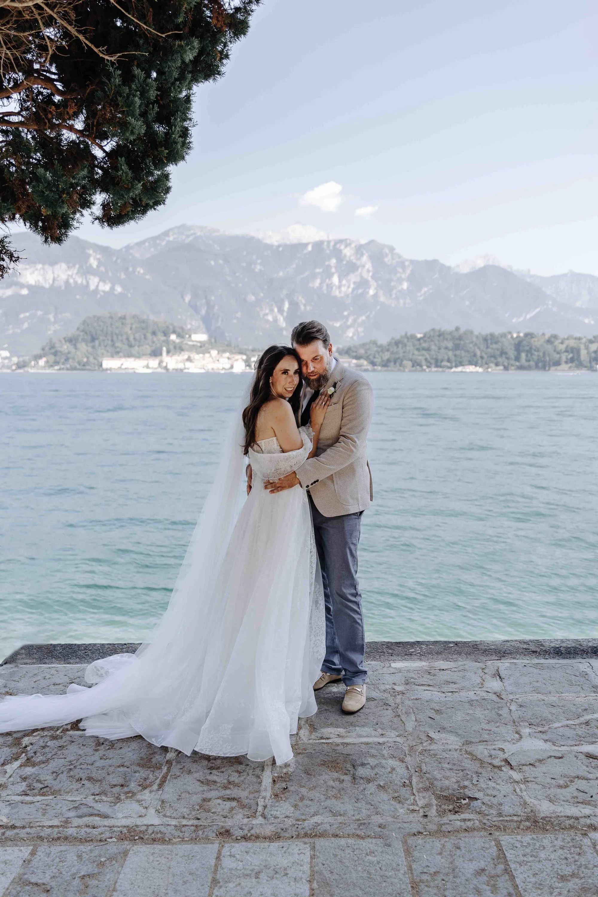 A bride and groom standing close together by a lake with mountains in the background, during daylight. The bride is in a white wedding dress with a long train, and the groom is in a light-colored suit. There is a large tree on the left side of the im
