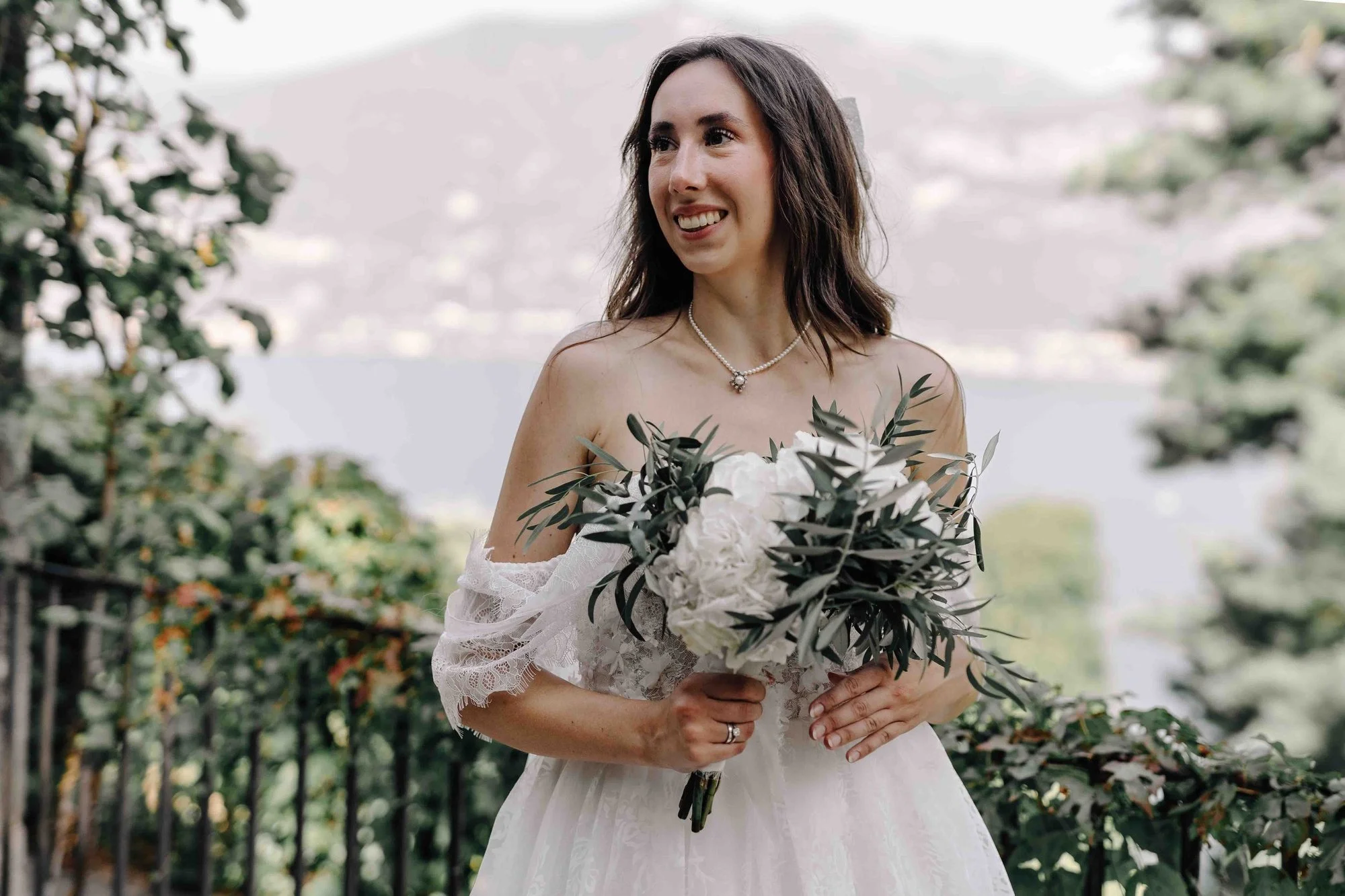 A woman in a wedding dress holding a bouquet of white flowers and green foliage outdoors, smiling, with trees and a body of water in the background.