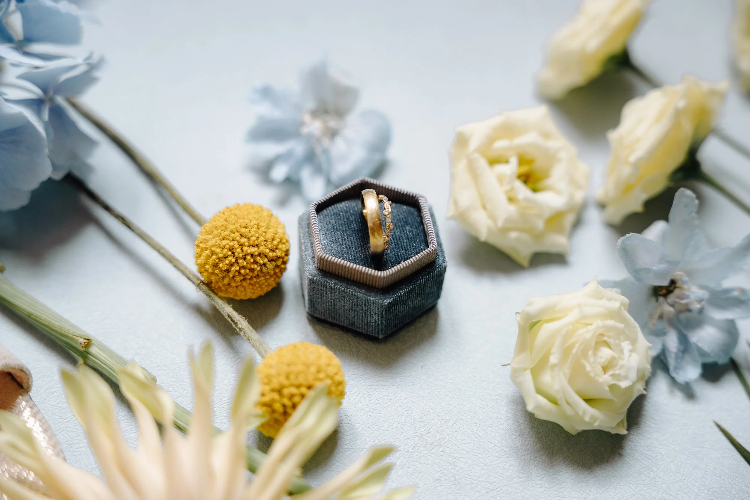 A gold ring with small diamonds in a blue jewelry box, surrounded by white and yellow flowers.