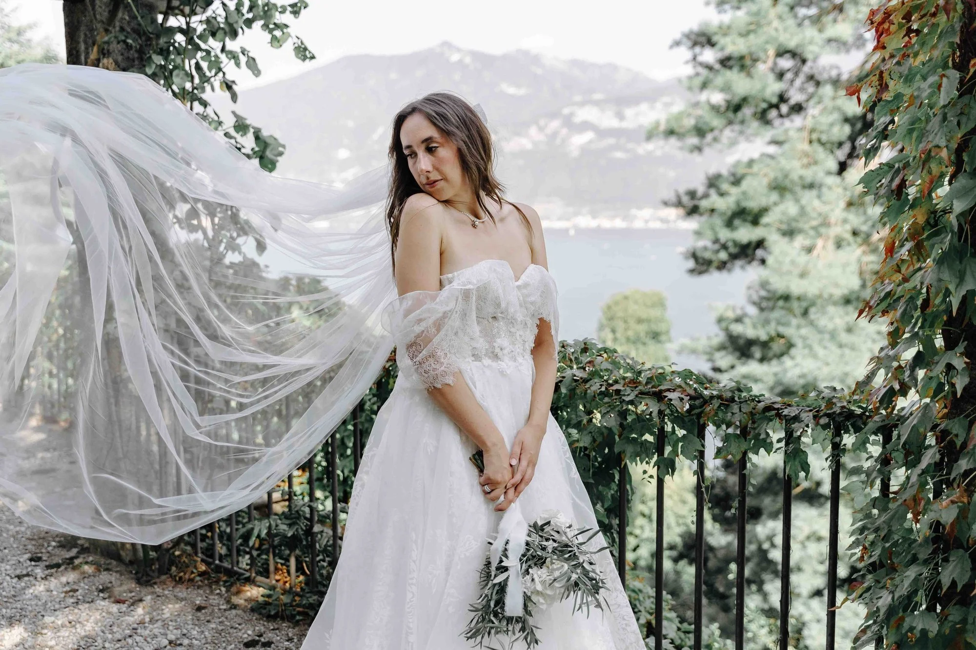 A woman in a white wedding dress and veil standing outdoors near a tree and foliage, with a lake and mountains in the background, holding a small bouquet of greenery.