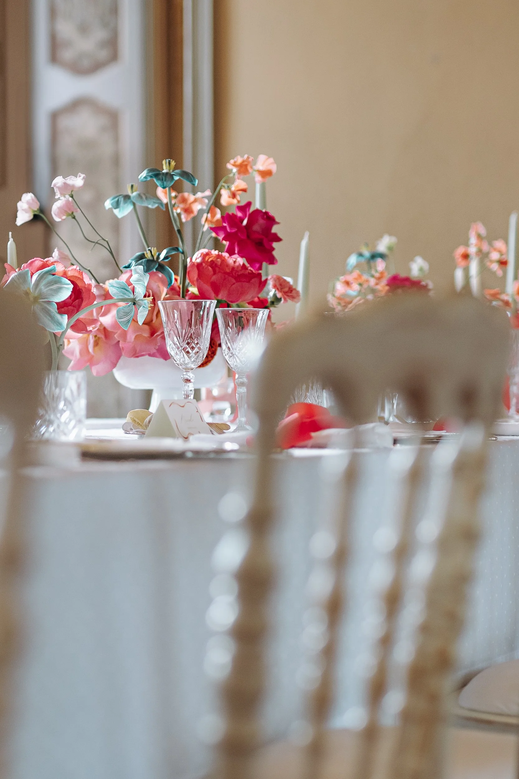 A table with pink, orange, and blue flower arrangements, crystal glassware, and place cards, set for a formal event or celebration.