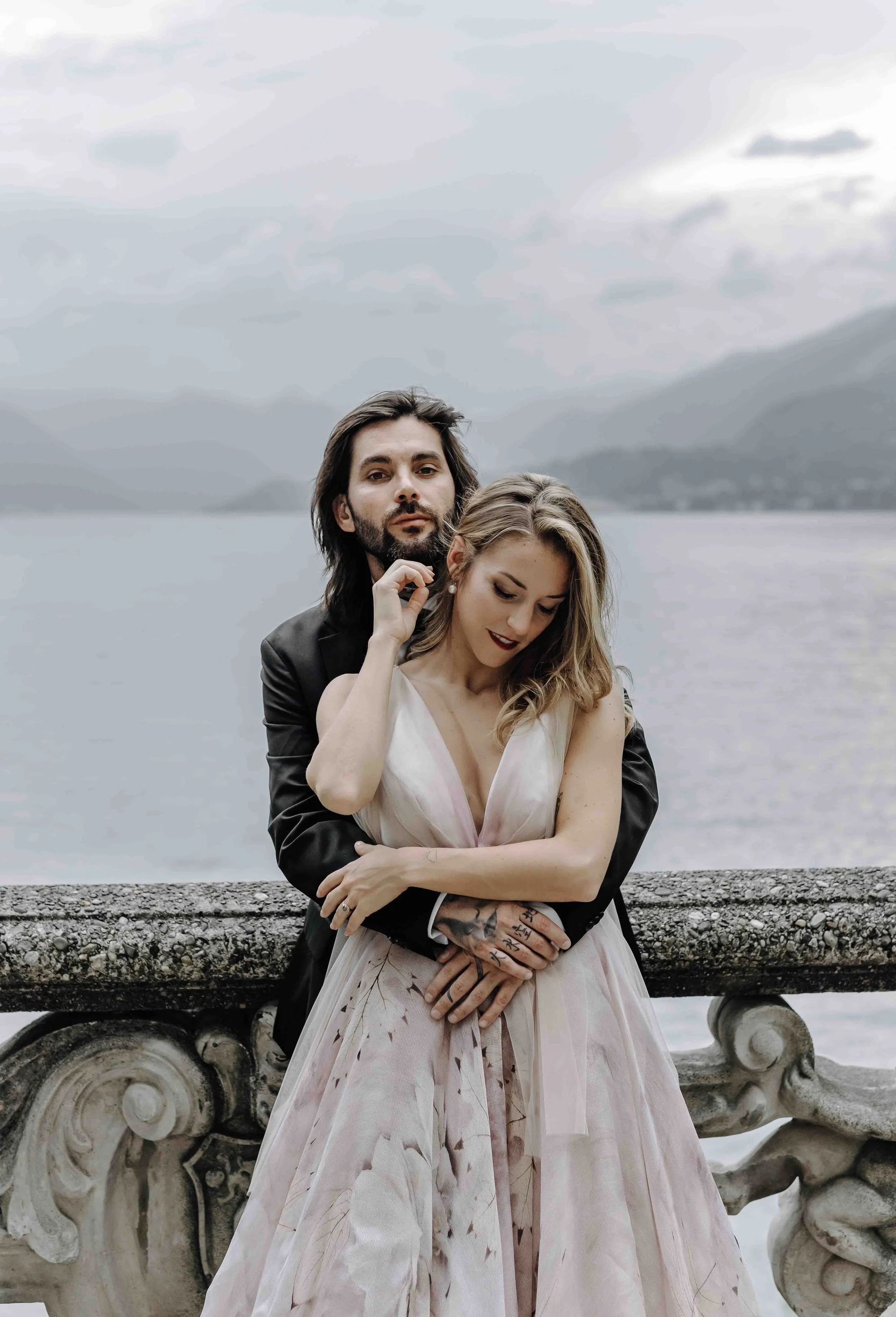 A couple stands close together outdoors by a stone railing, with mountains and a lake in the background. The man has long dark hair, a beard, and is wearing a black jacket. The woman has long blonde hair, is wearing makeup, and a light-colored elegan