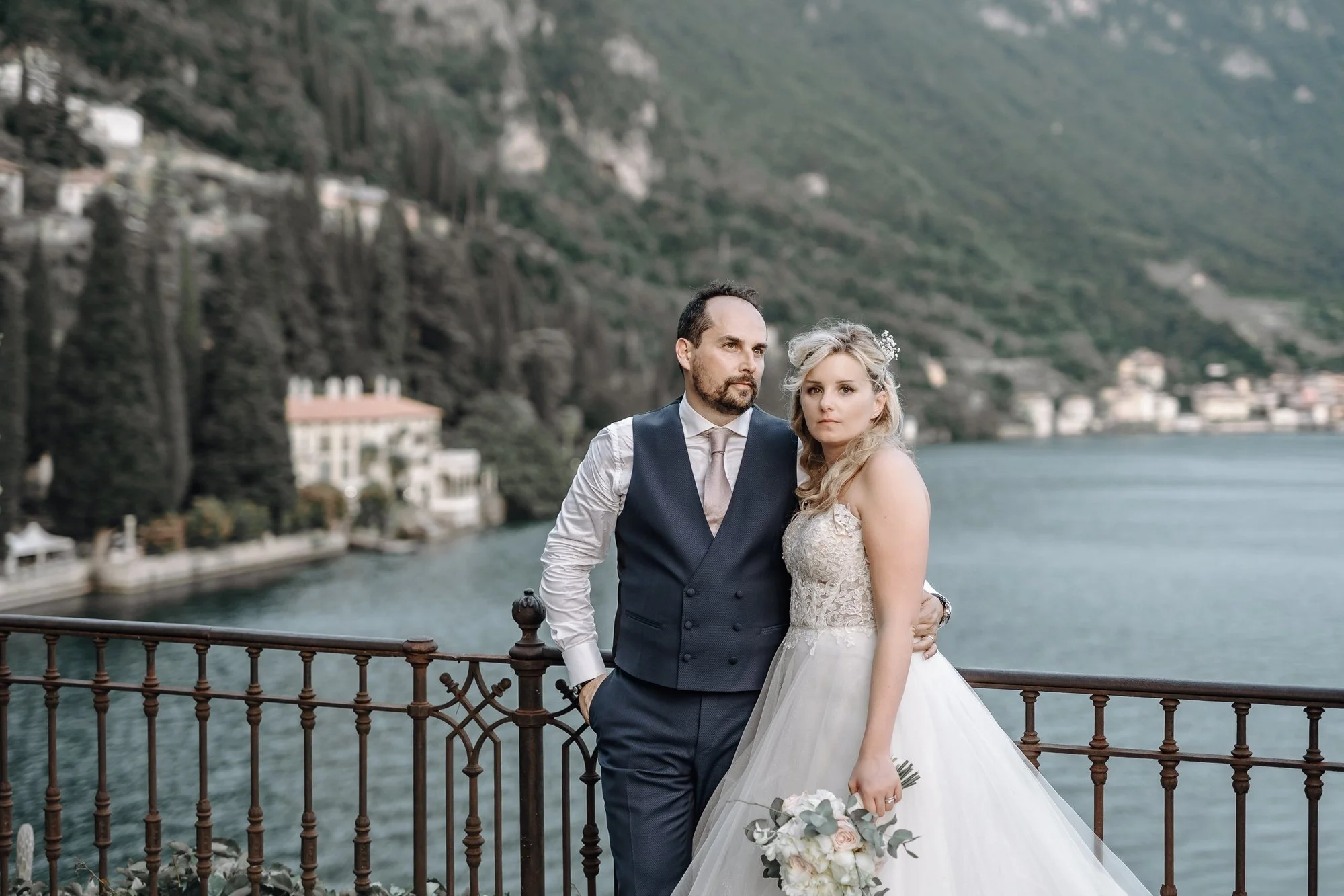 A bride and groom standing together outdoors by a lake with mountains in the background, dressed in wedding attire, with the bride holding a bouquet and the groom with his arm around her.