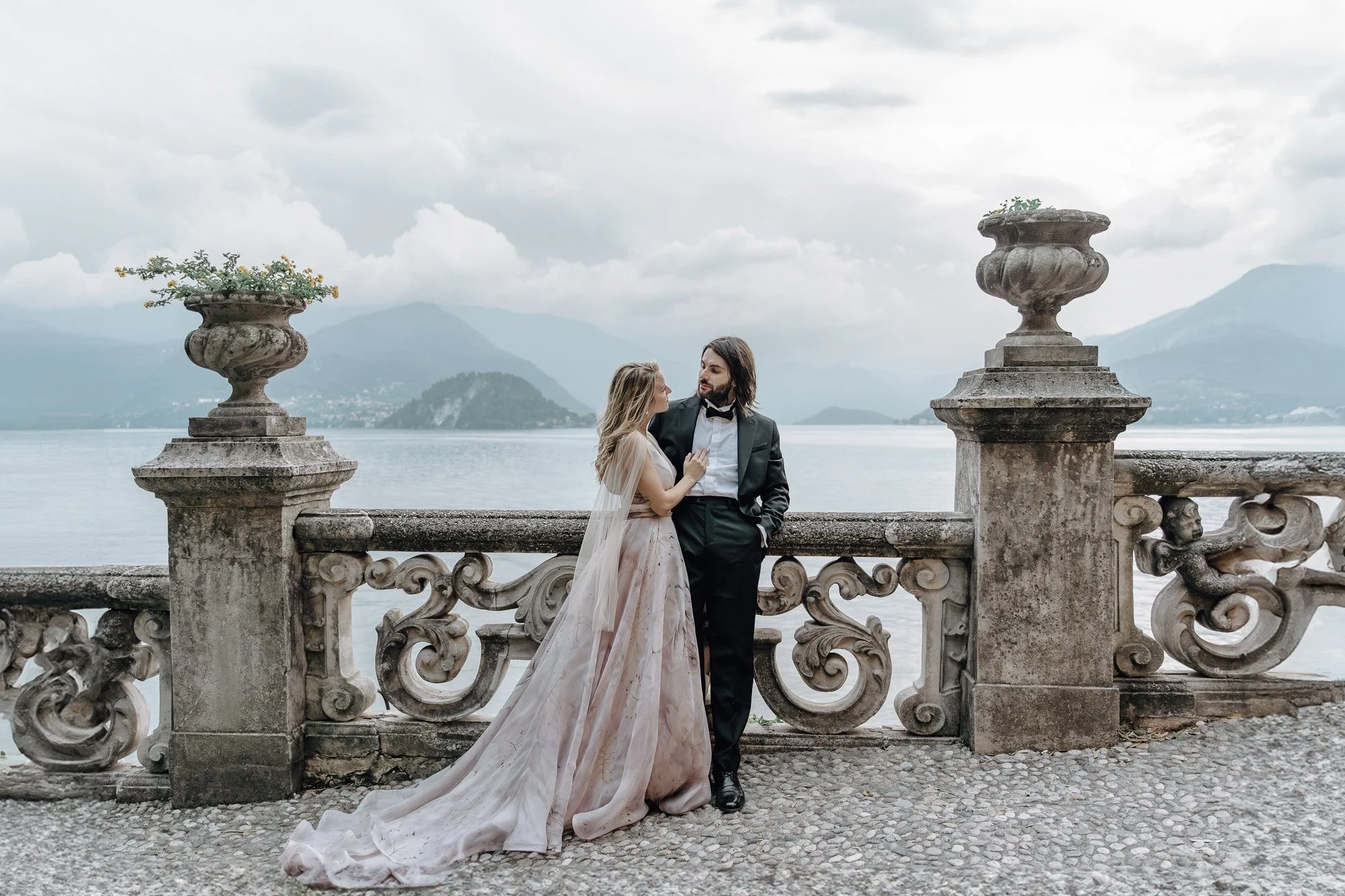 A couple dressed in wedding attire standing on a stone balcony overlooking a lake with mountains in the background. The woman is wearing a long, flowing gown and the man is in a tuxedo. The scene is romantic and scenic with cloudy skies.