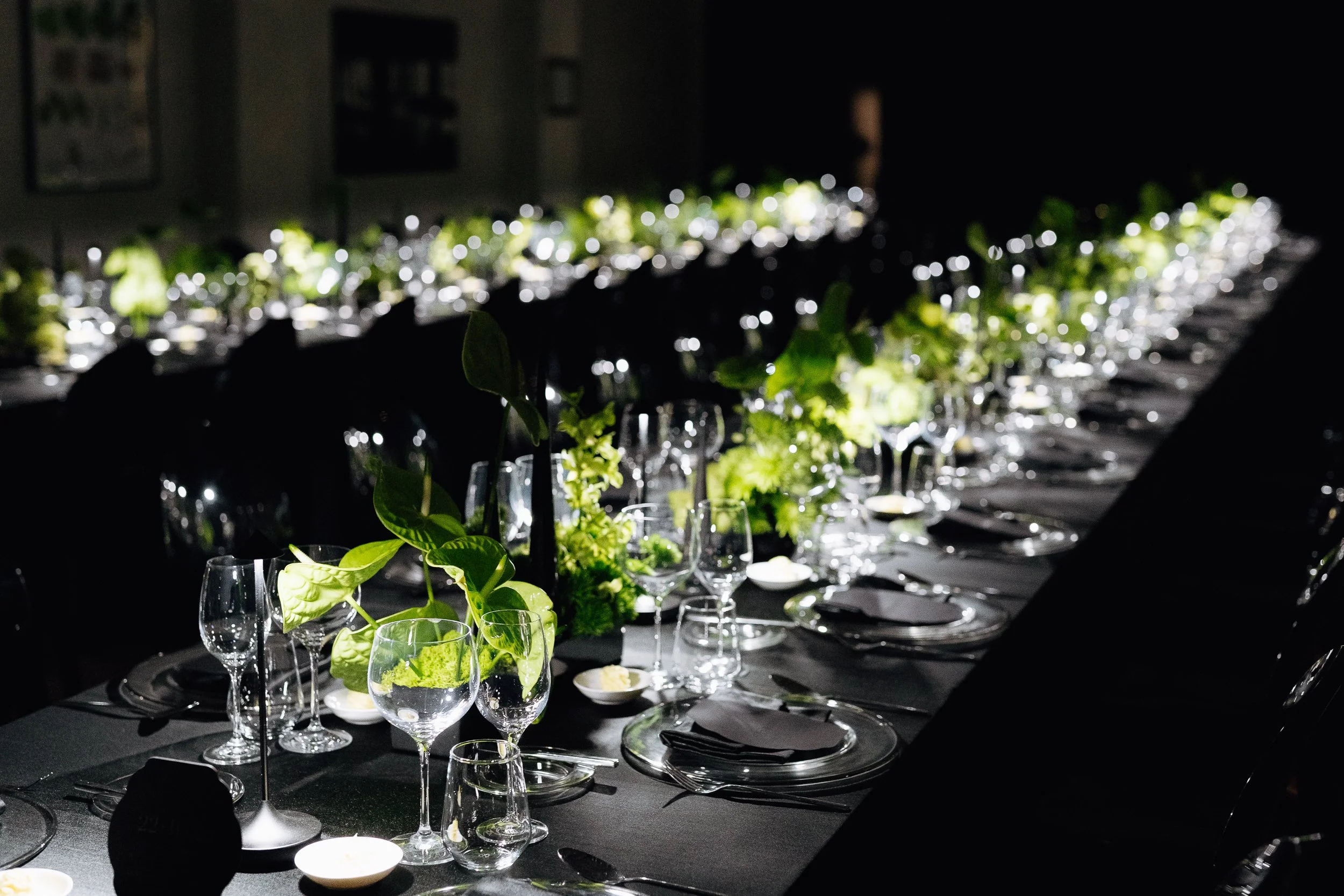 A long dining table set for an event or celebration, decorated with green plants and foliage, with glassware and plates arranged neatly. The room is dimly lit with a focus on the table decorations.