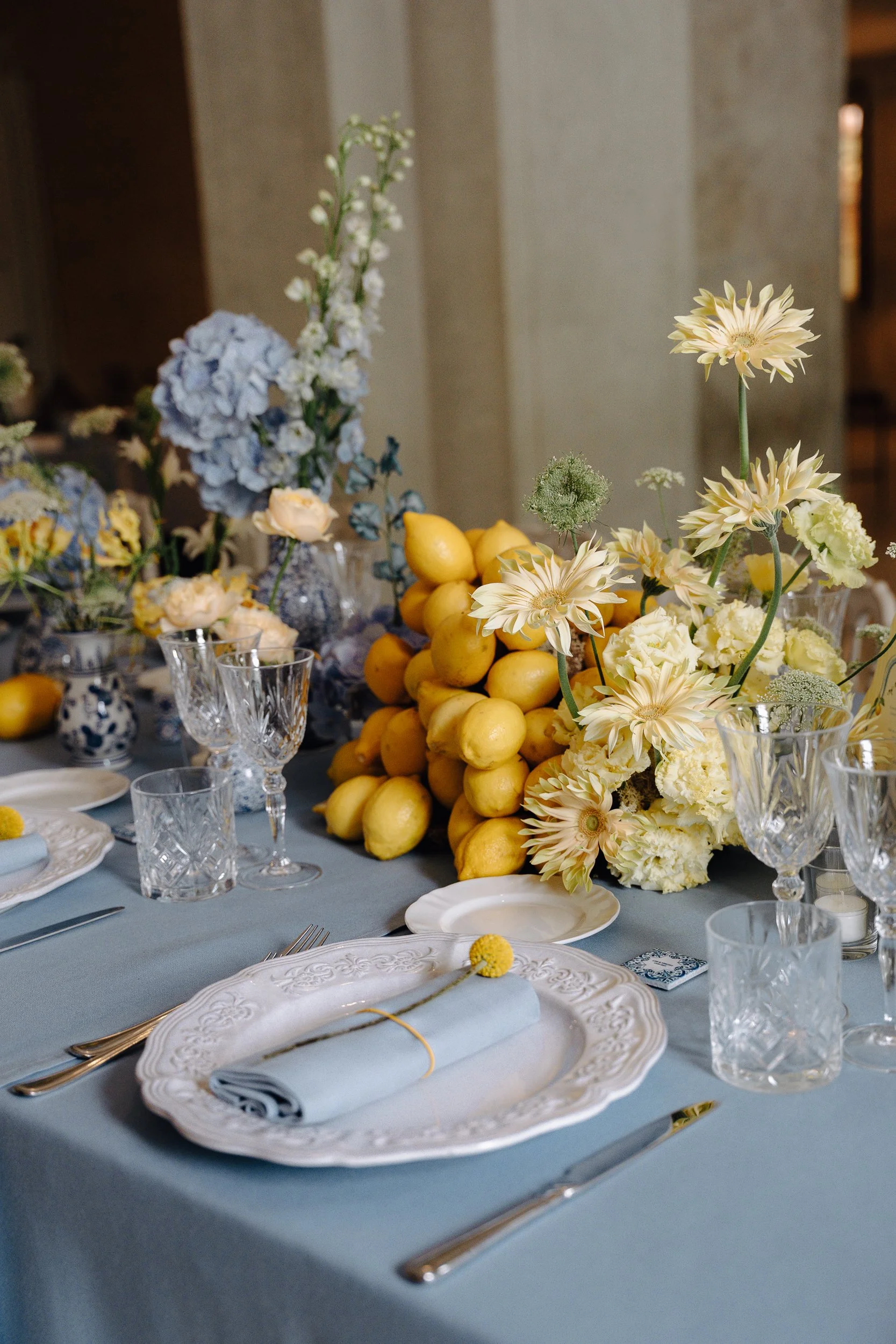 A table decorated for a formal event with white and cream floral centerpieces, lemons, and elegant place settings that include white plates, silverware, clear glassware, and light blue napkins.