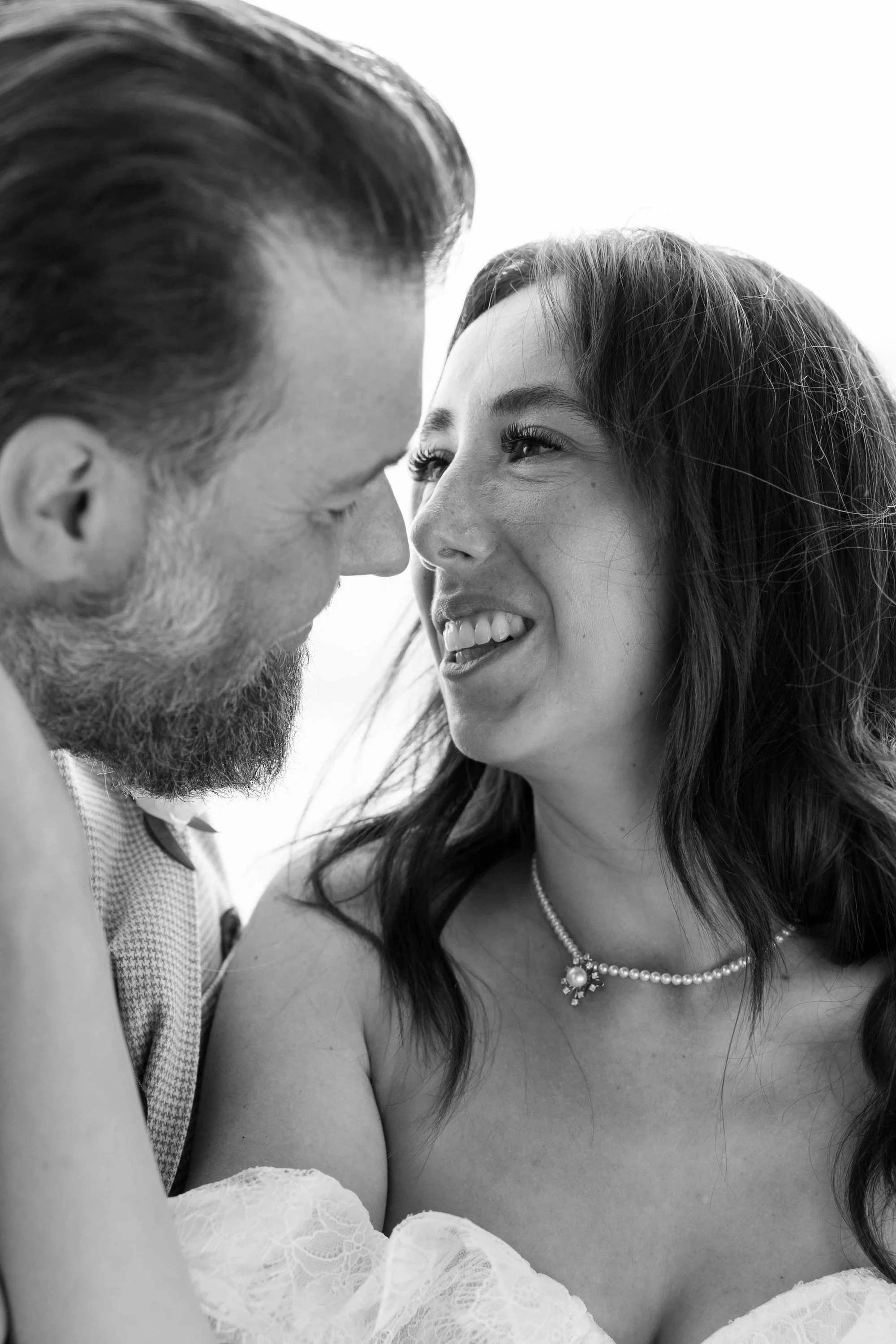 A black and white close-up photo of a smiling woman wearing a pearl necklace, an ornate brooch, and lace off-the-shoulder dress, with a man whose face is close to hers, both smiling warmly.