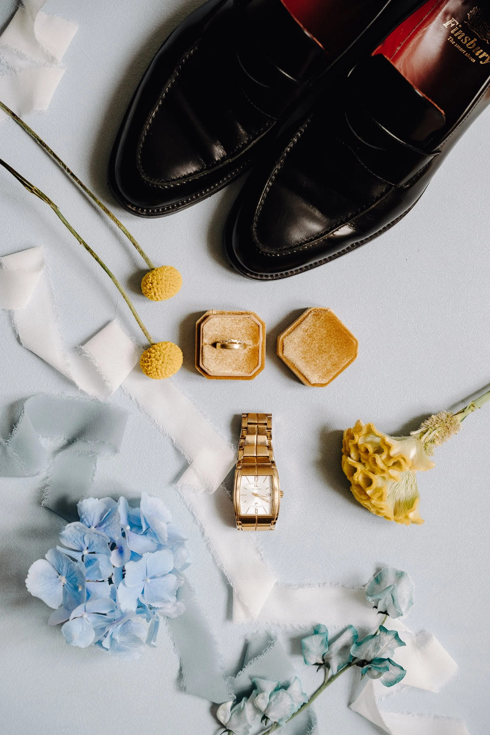 A flat lay of black leather shoes, a gold wristwatch, a wedding ring in a velvet box, dried flowers, and decorative paper ribbons on a white surface.