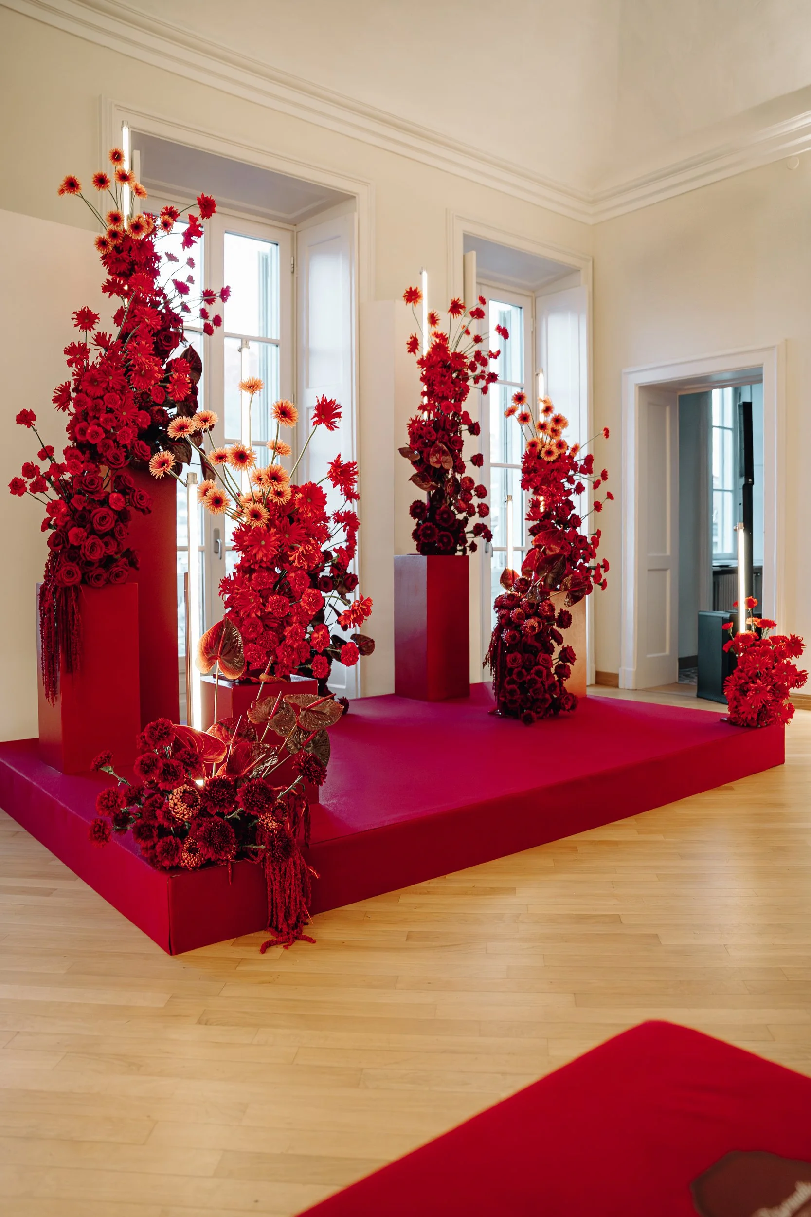 Indoor art installation with red flower arrangements on red pedestals in front of large windows in a bright room.