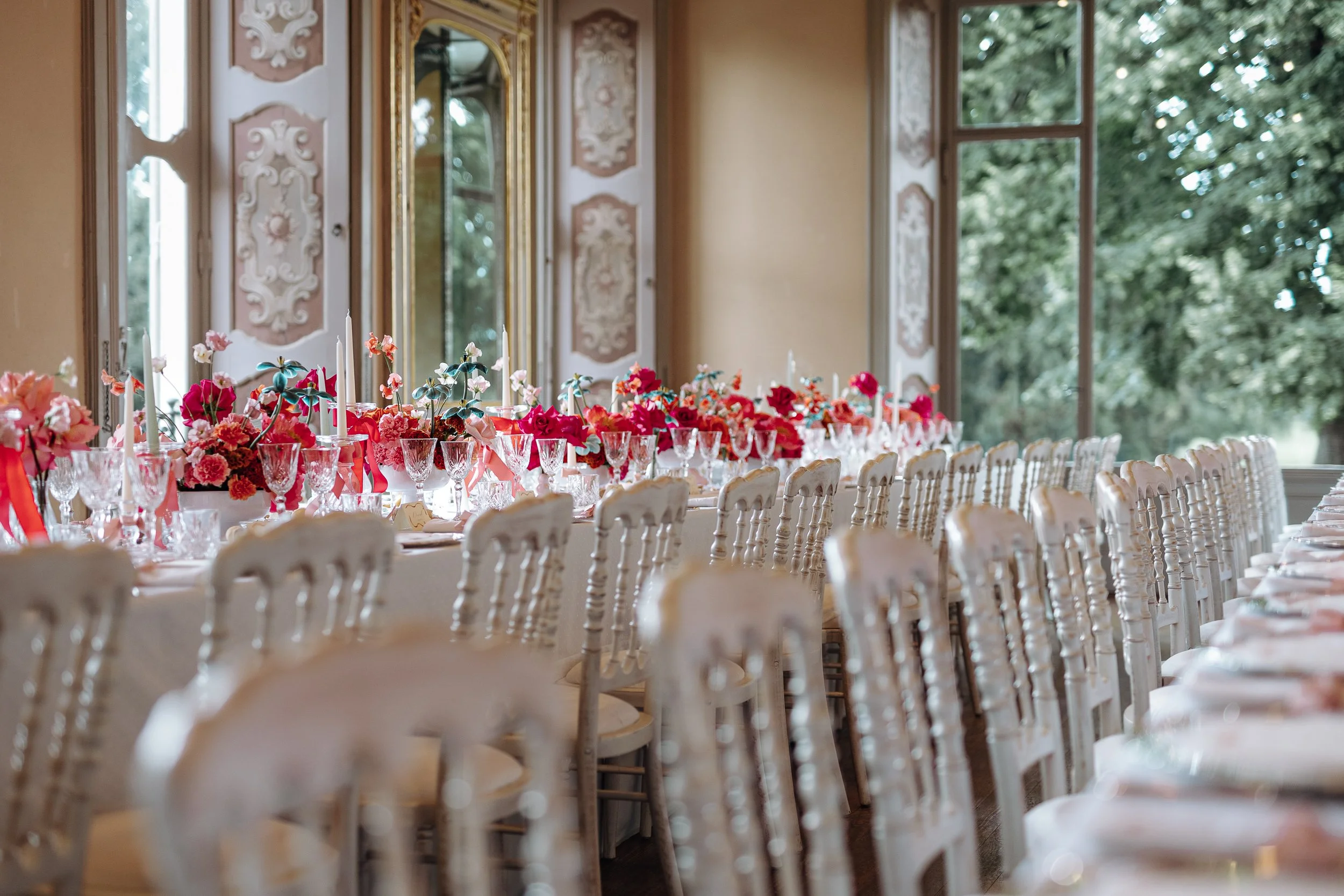 A long banqueting table decorated with pink and red flowers, candles, and glassware, set in a room with large windows and ornate wall decor.