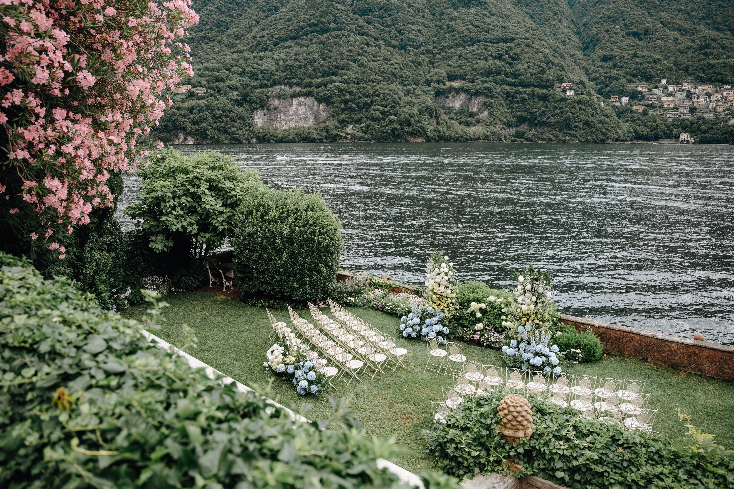 Outdoor lakeside wedding setup with white chairs arranged in rows on green grass, surrounded by flower arrangements and lush green foliage, overlooking a river with a forested hillside in the background.