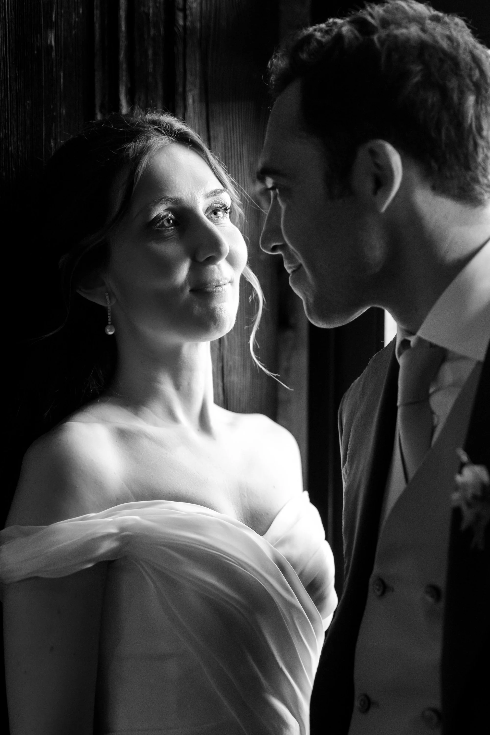 A black-and-white photo of a woman in an off-shoulder dress and pearl earrings looking at a man in a tuxedo with a boutonnière, standing close together in front of a wooden wall.