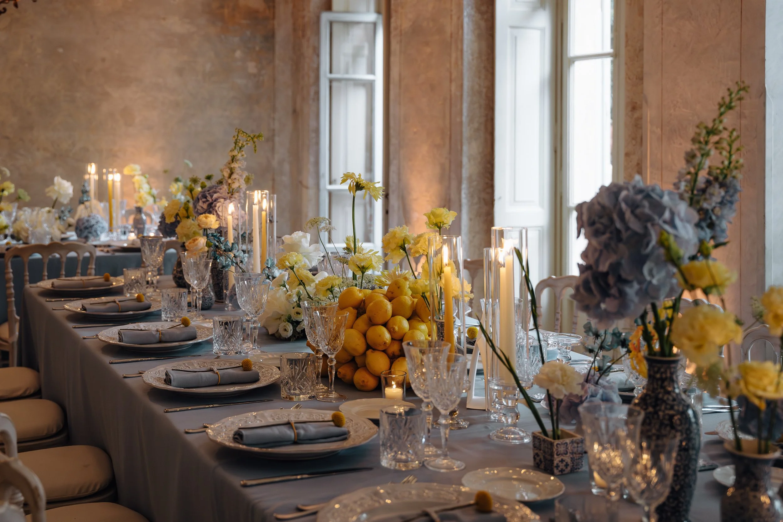 Elegant dining table decorated with yellow and white flowers, candles, and lemons in a bright room with tall windows.