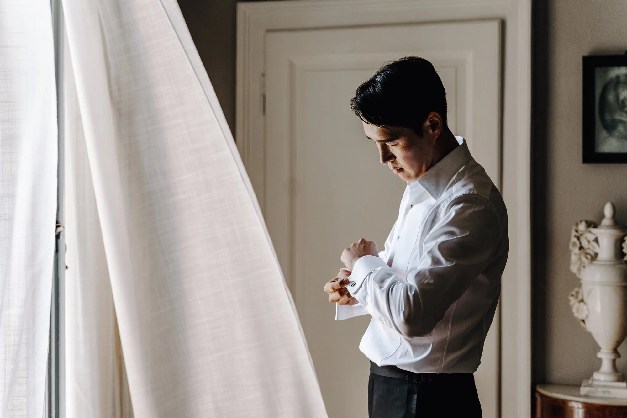 A man in a white dress shirt is standing near a window, adjusting his cufflink. The scene is indoors with natural light coming through the window, and a door and decorative items are visible in the background.