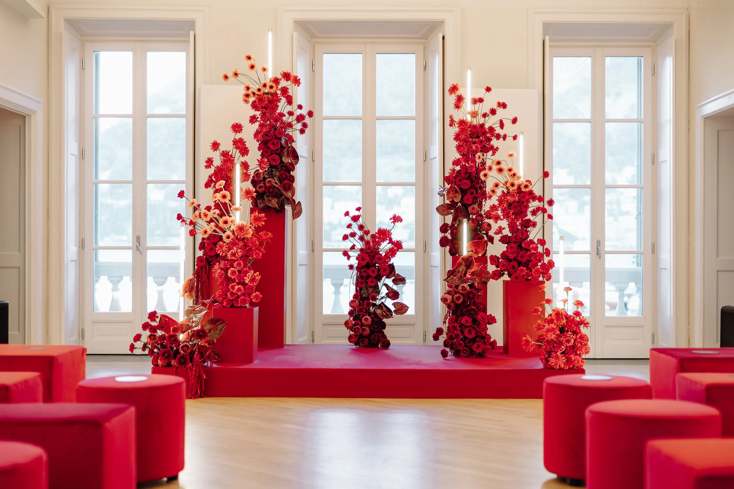 An indoor room with a large windowed wall, decorated with multiple tall, red floral arrangements on red pedestals, and red cushioned seats in the foreground.