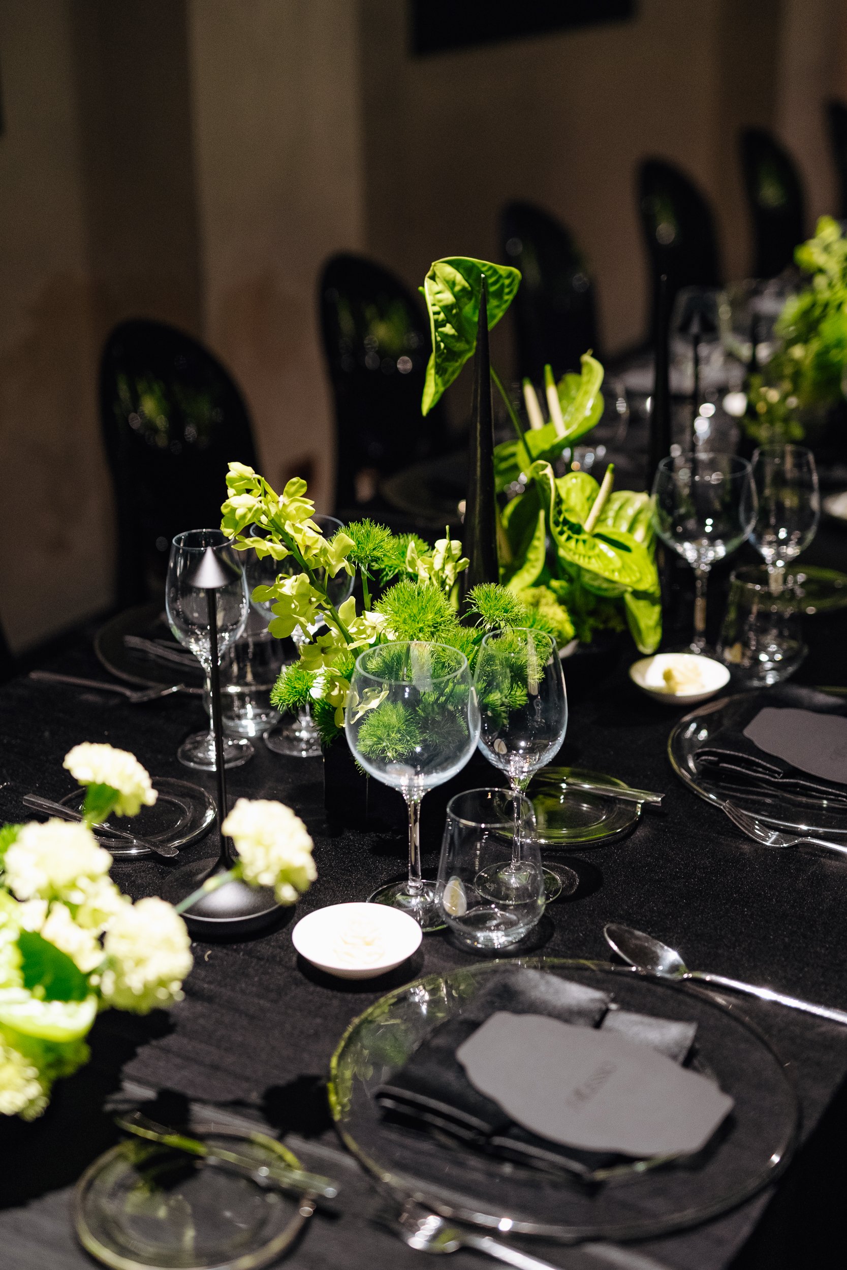 Elegant dinner table setting with glassware, black tablecloth, and floral centerpiece with green leaves and white flowers.
