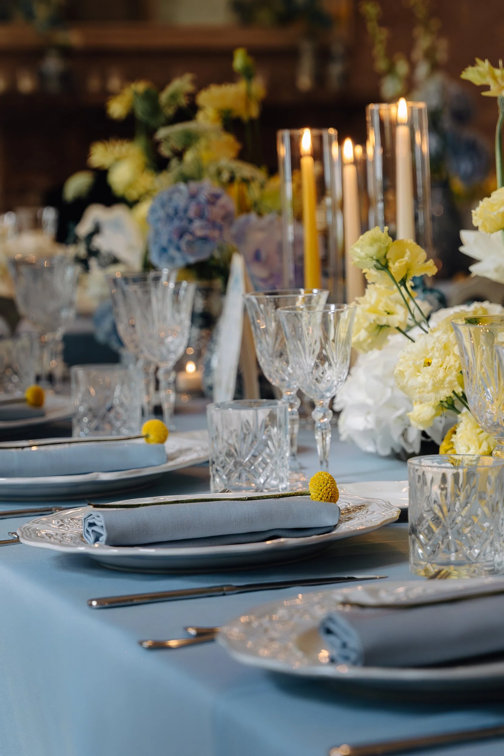 Elegant table setting with white and blue linens, crystal glassware, silverware, and floral centerpieces featuring white and yellow flowers, with tall candles.