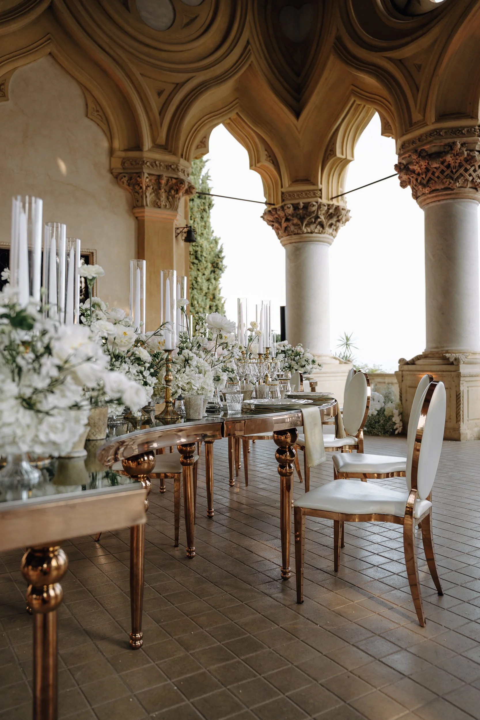 Elegant round dining table set with white flowers, candles, and glassware, situated on a tiled floor beneath a detailed, arched ceiling supported by large columns, with a view of trees and bright sky outside.