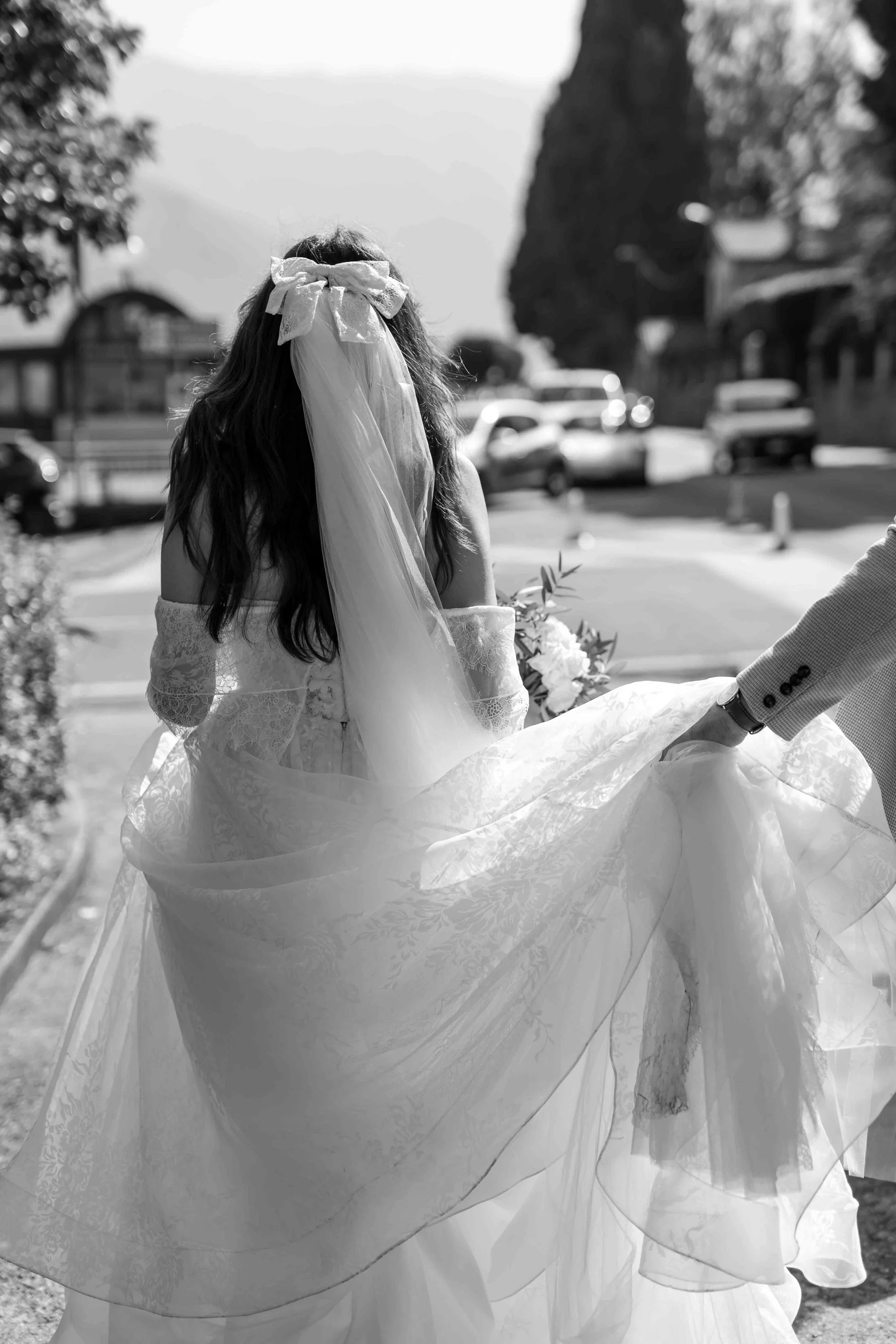 A woman wearing a wedding dress holding a bouquet, with her back to the camera, outdoors on a sunny day.