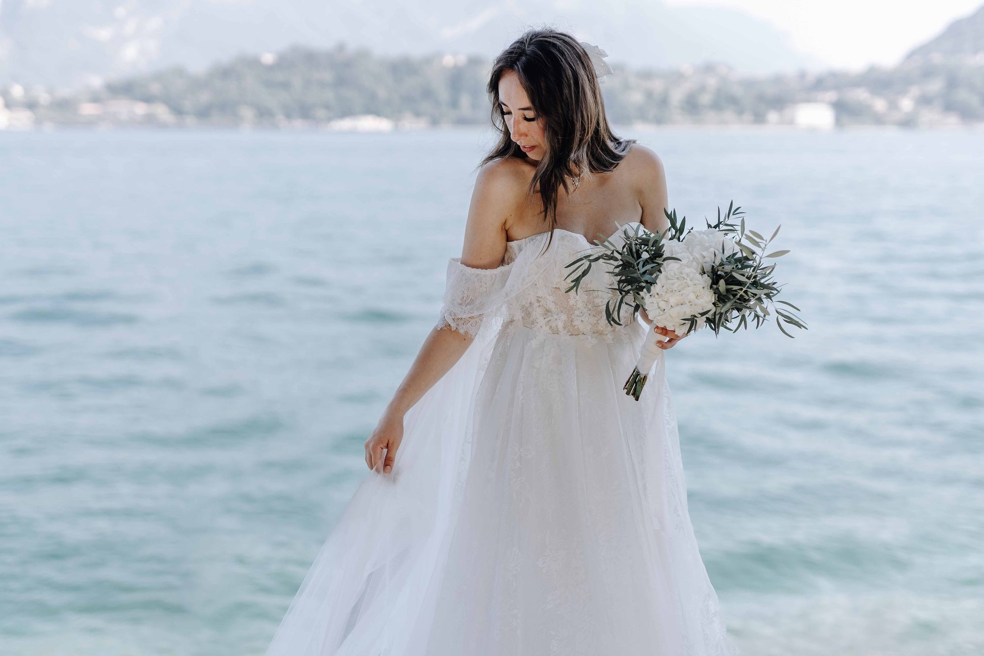 A woman in a white wedding dress holding a bouquet of white flowers with greenery, standing near the water with a blurred background of a distant shoreline and hills.